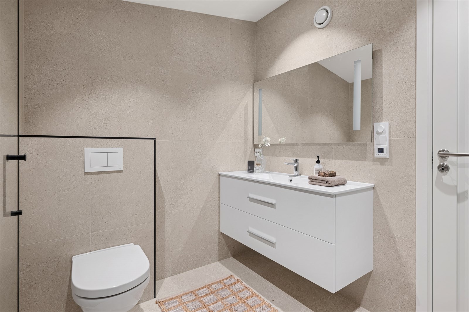 Elegant baderom med beige fliser, vegghengt toalett, servant, og speil med innebygde LED-lys. Galleribilde