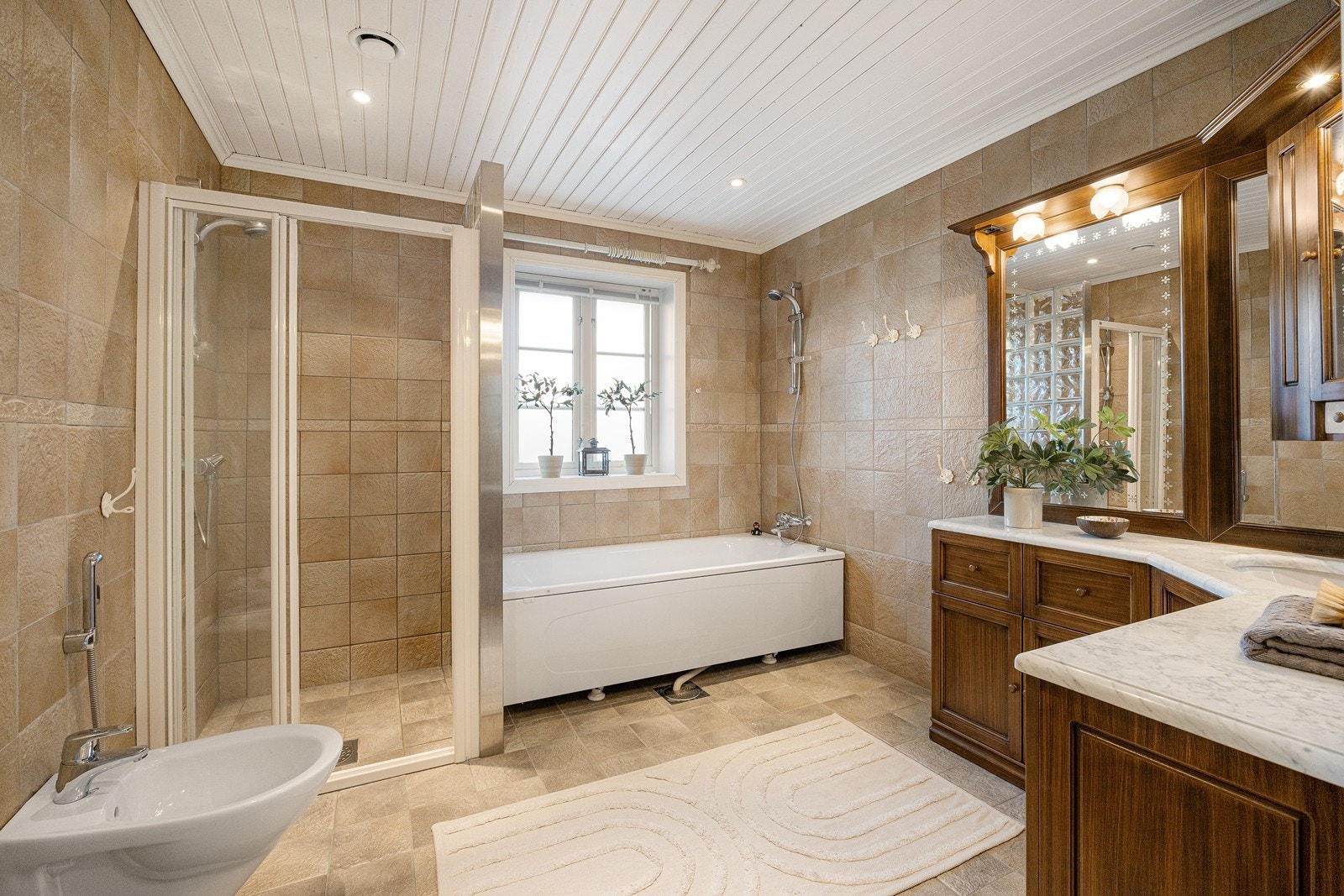 Badet er av god størrelse, delikat innredet med grå vegg- og gulvfliser. En vegg i glassbyggerstein skiller et badekar med dusjløsning og et dusjhjørne med skyvedør av glass. Galleribilde