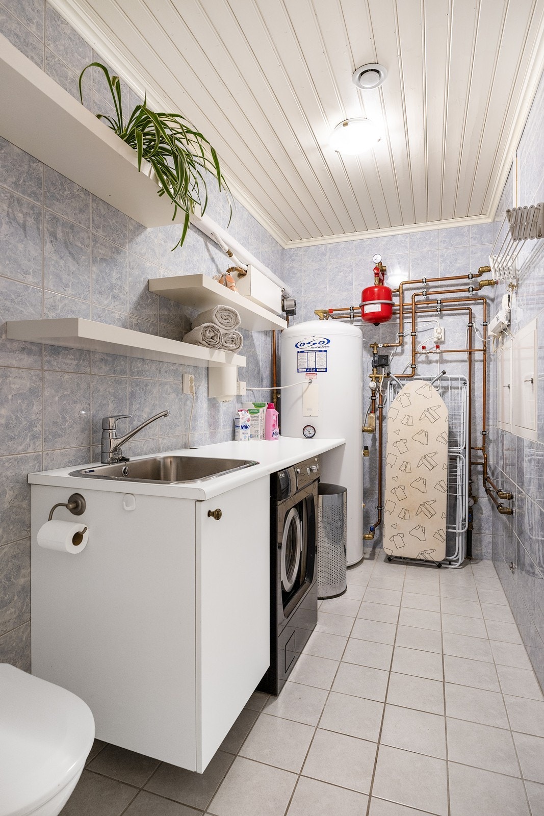 Vaskerommet har flislagt gulv, åpne hyller, toalett og arbeidsbenk med nedfelt utslagsvask. Det er opplegg for vaskemaskin og plass til tørketrommel. Galleribilde