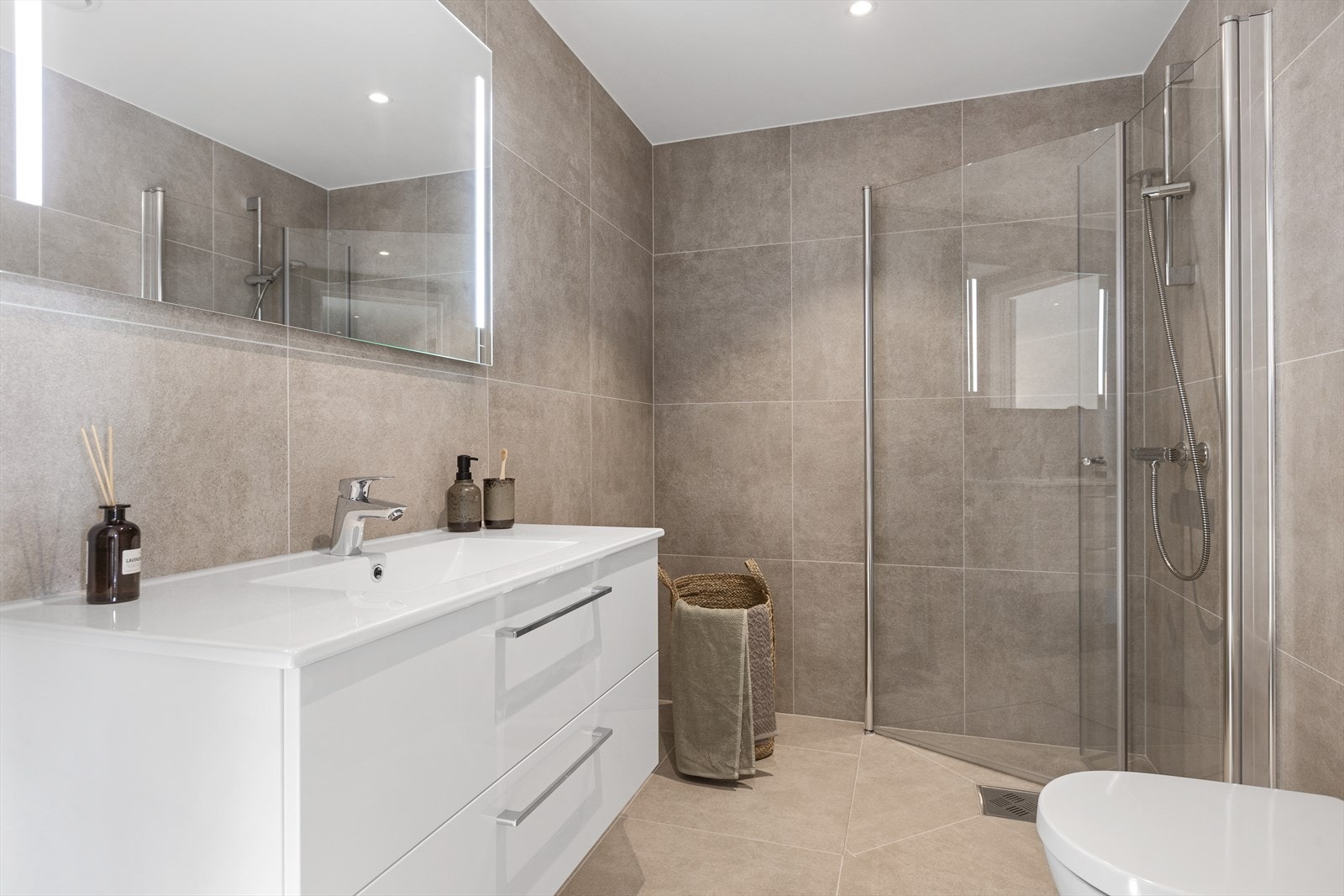 Lekkert Bad/wc med flislagte vegger og flislagt gulv med vannbåren gulvvarme. Malt gipstak med innfelte LED downlights med dimmer. Veggmontert wc. Dusjhjørne med 90x90 cm glassvegger. Slett, stilren baderomsinnredning med LED speil Galleribilde