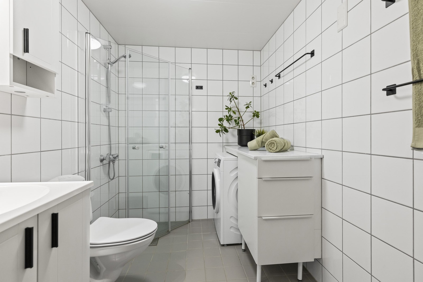 Badet består av vegghengt servantinnredning, heldekkende servant, vegghengt skap over servant med overlys og stikkontakt, gulvstående benk med benkeplate, dusjhjørne med innfellbare glassdører, dusjarmatur tilkoblet hånddusj og vegghengt toalett Galleribilde