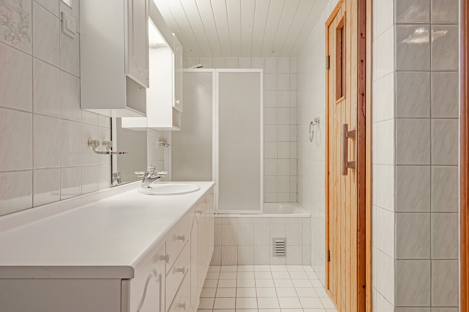 Flislagt baderom i kjelleren med badekar med vegg og dusj, servant på innredning og skap med speil. Galleribilde