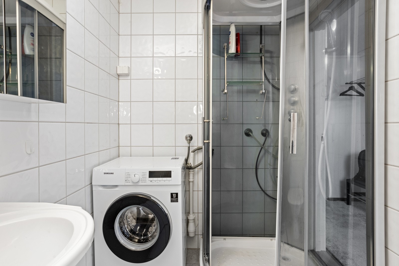 Baderom med vegghengt servantinnredning, speilskap med overlys og stikkontakt, dusjkabinett og opplegg for vaskemaskin Galleribilde