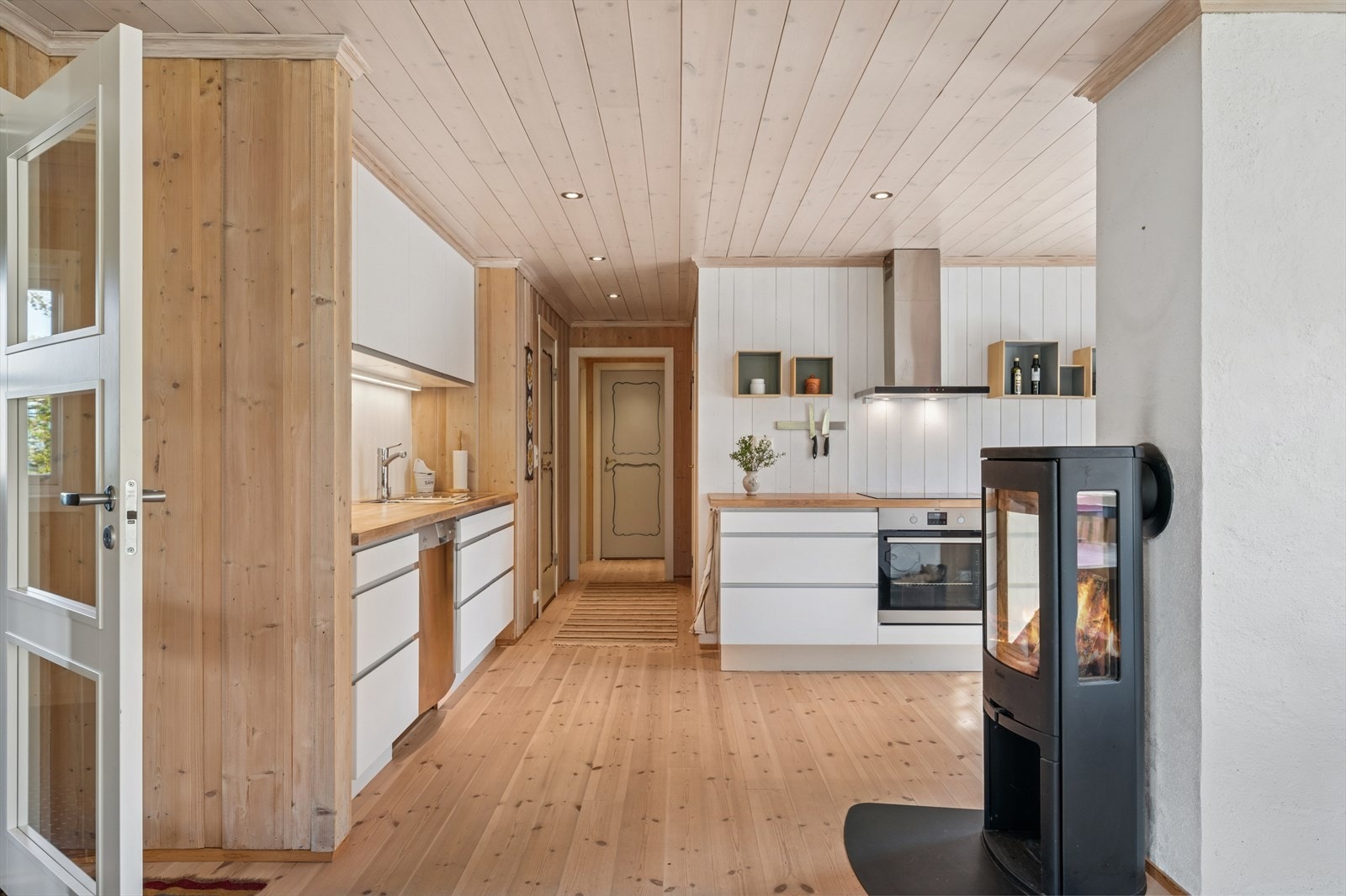 Videre kommer vi til det lyse og moderne kjøkkenet med hvite slette fronter og integrerte hvitevarer som gir et helhetlig og stilrent uttrykk. I tilknytning til kjøkkenet finner vi også en koselig peis - perfekt for å skape varme og stemning. Galleribilde