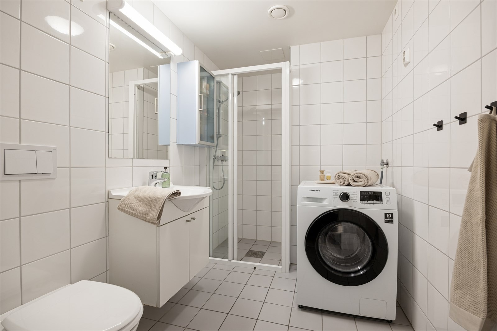 Flislagt baderom med praktisk løsning, utstyrt med servant nedfelt i skapinnredning, wc og dusjhjørne. Opplegg for vaskemaskin. Galleribilde