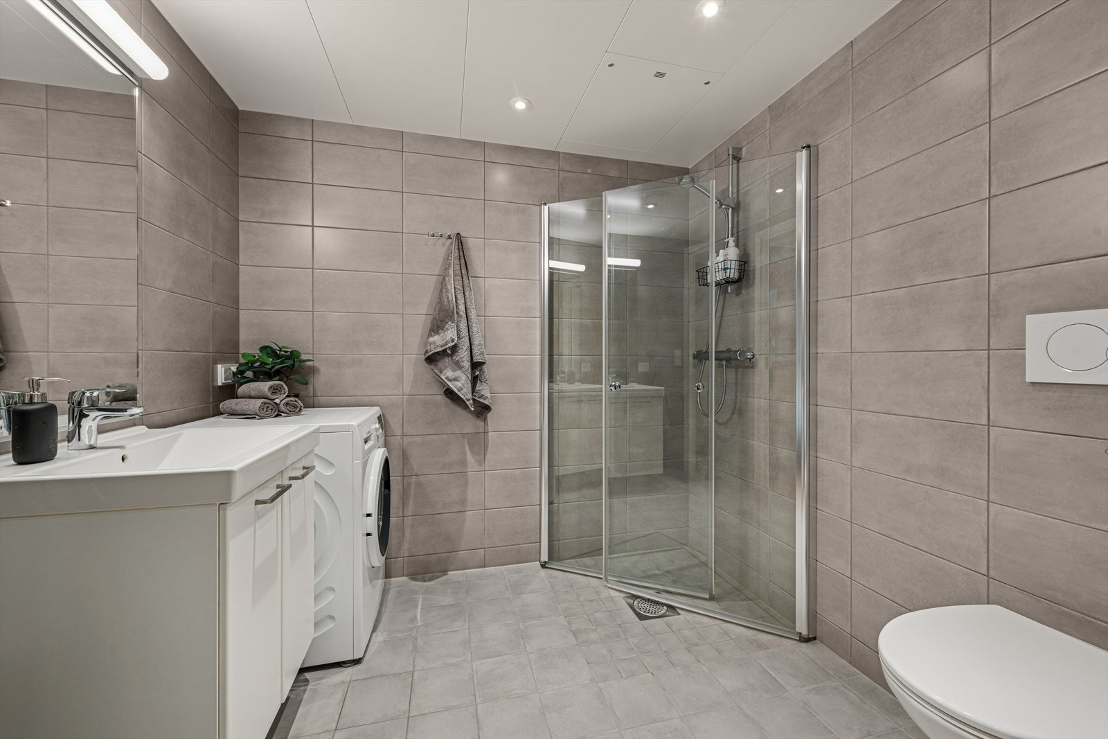 Badet er innredet med vegghengt toalett, servant med underskap, innfellbare dusjvegger og opplegg til vaskemaskin. Galleribilde