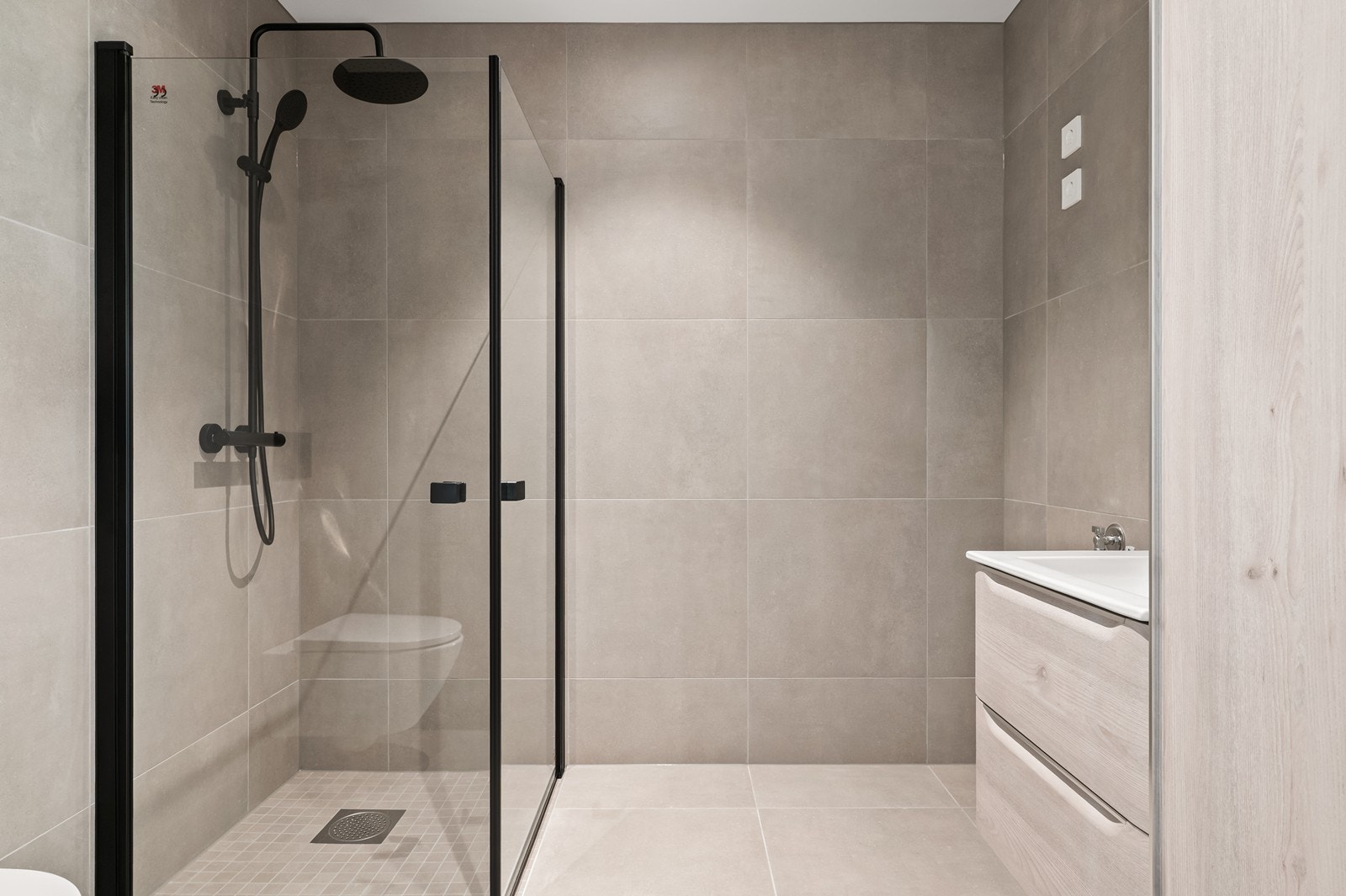 Delikat, flislagt bad med vegghengt toalett og dusjhjørne med sorte profiler. Galleribilde