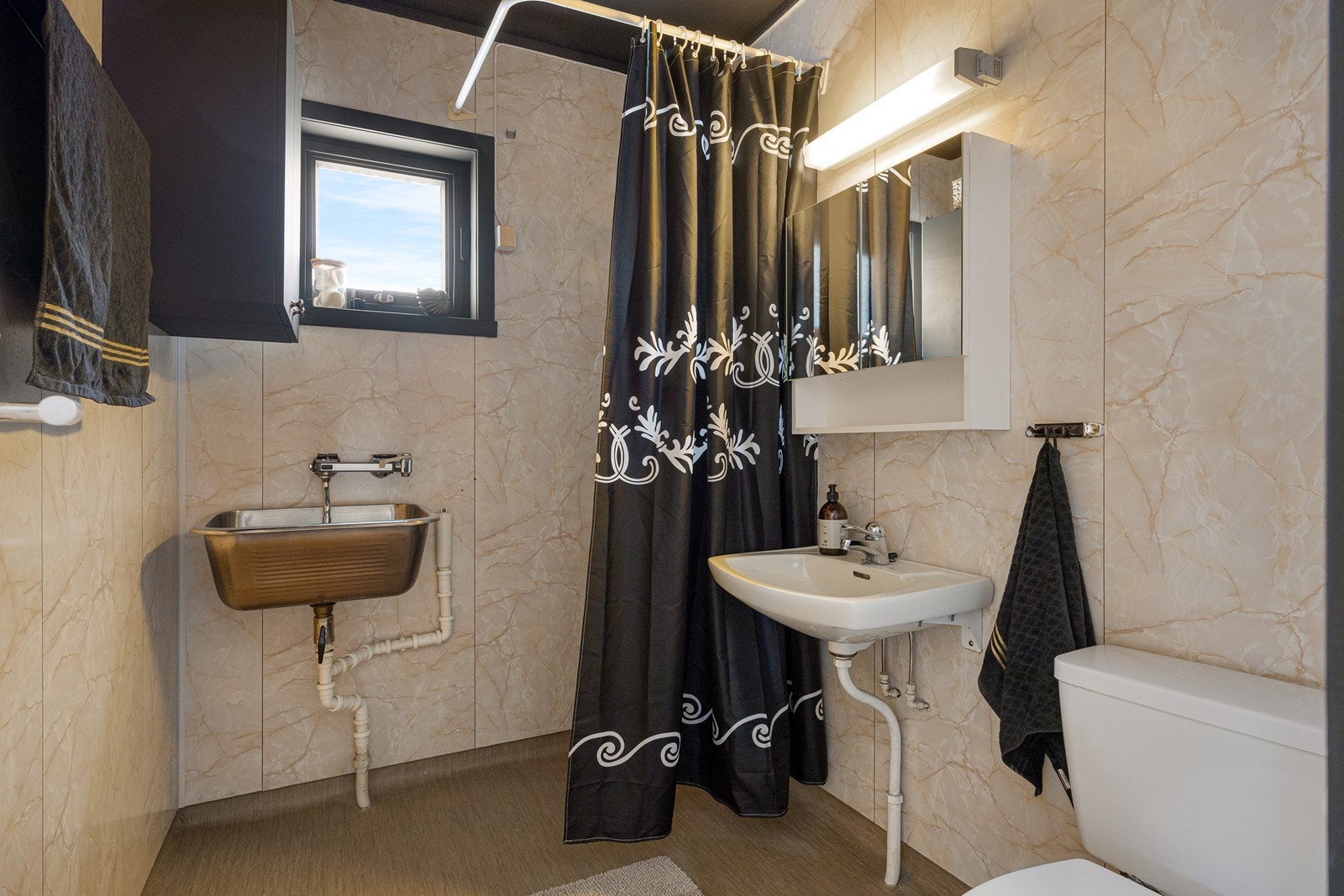 Badet i 1. etasje har lyse våtromsplater og grått belegg på gulv med sluk. Badet er videre utstyrt med et toalett, dusjarmatur, utslagsvask og opplegg for vaskemaskin. Galleribilde