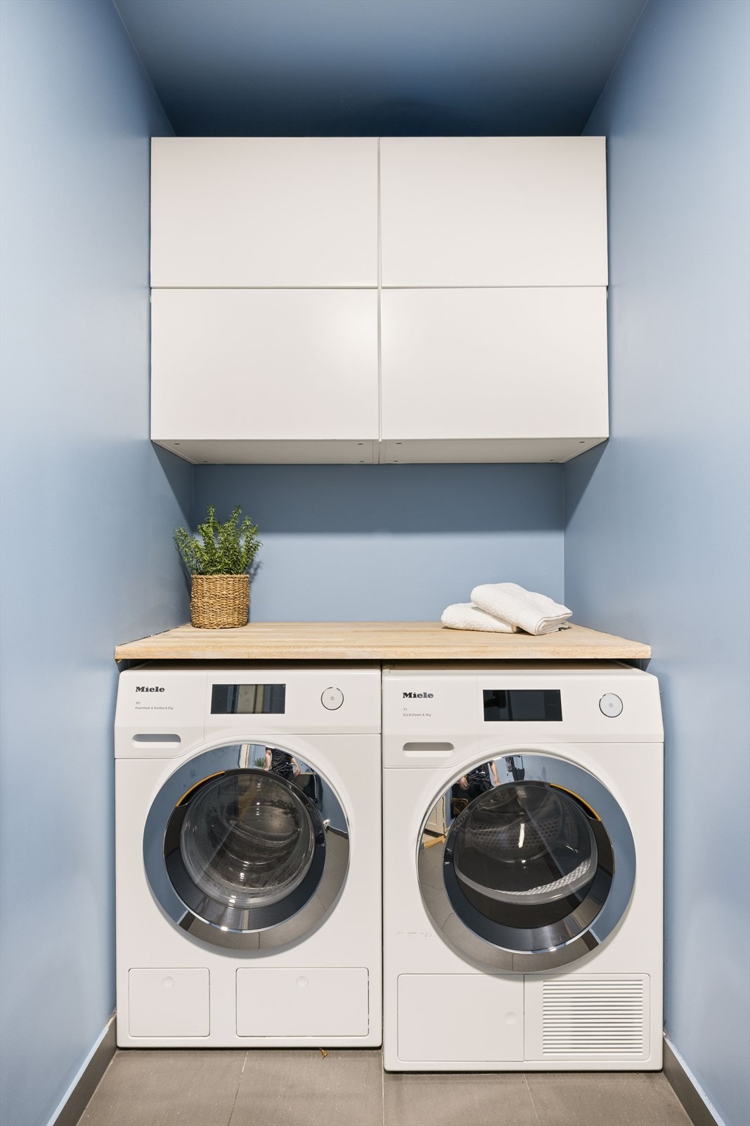Vaskerommet har store, grå gulvfliser med varme, mens både tak og vegger er malt i en moderne blåfarge. Plassen er godt utnyttet, med hvite overskap og to benkeplater i heltre. Galleribilde