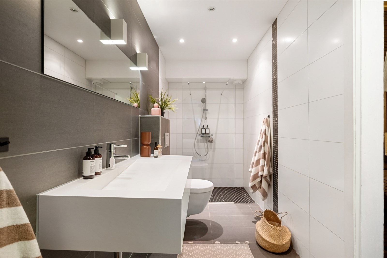 Badet er utstyrt med vegghengt servant med ettgreps blandebatteri, vegghengt speil og veggmontert toalett fra Geberit. Den romslige dusjsonen (ca. 150 x 90 cm) har fastmontert dusjvegg i herdet glass. Galleribilde