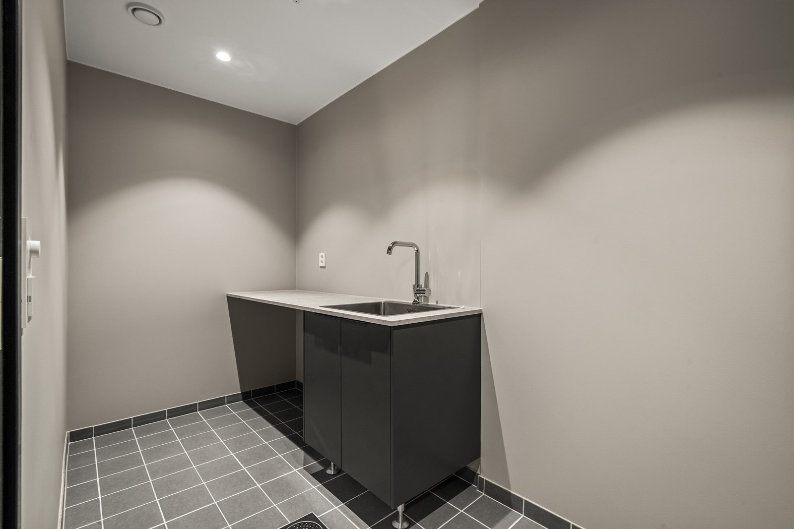 Praktisk vaskerom i kjelleretasje med flislagt gulv og malte veggflater. Praktisk skapinnredning med oppbevaring, benkeplate og oppvaskkum Galleribilde