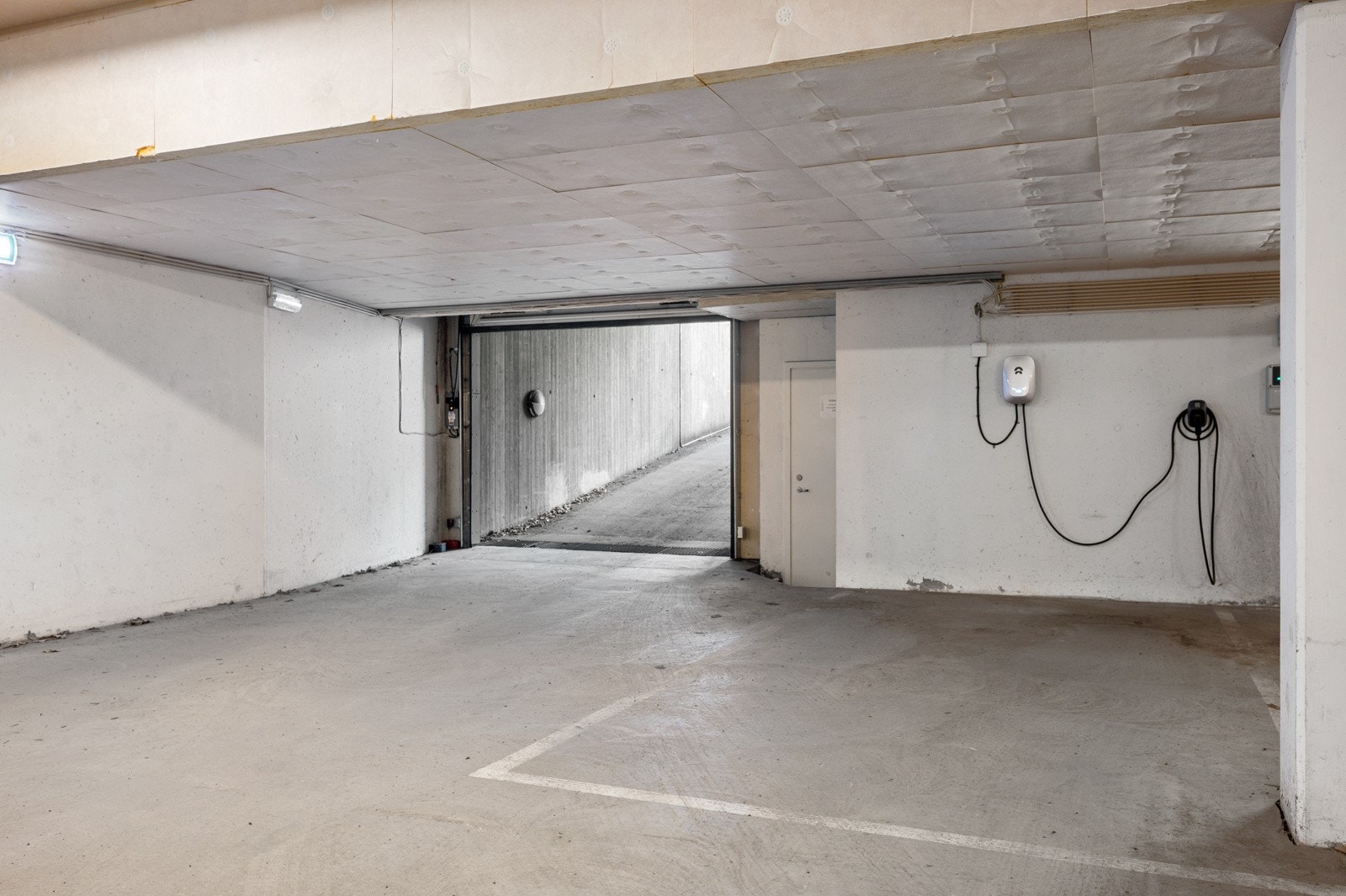 To parkeringsplasser i felles, lukket garasjeanlegg under bebyggelsen. Det er montert elbillader på den ene plassen. Galleribilde
