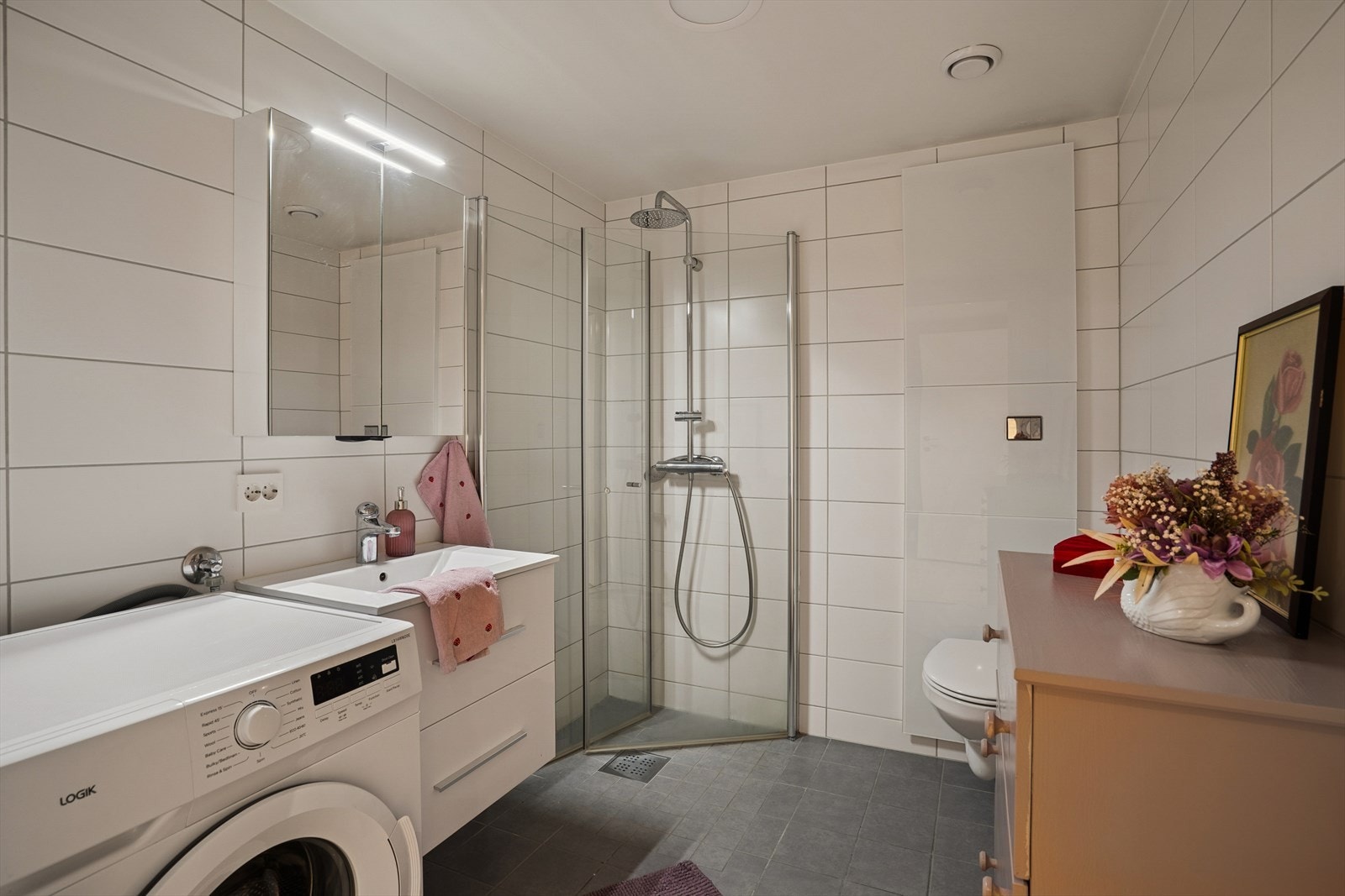 Badet er utstyrt med vegghengt toalett, dusjhjørne med innfellbare glassdører, baderomsinnredning og opplegg for vaskmaskin Galleribilde