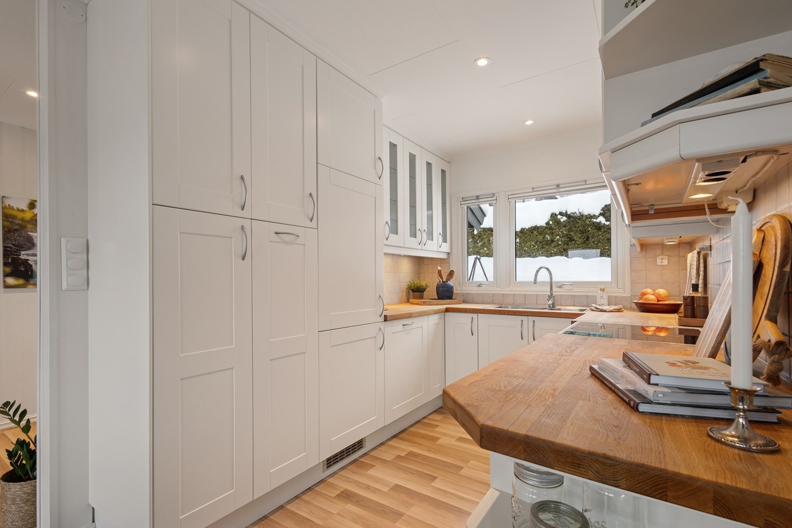 Innredningen har profilerte, hvite fronter og heltre benkeplate med dobbel kjøkkenkum. Det er flis over benkeplate og belysning under overskap. Galleribilde