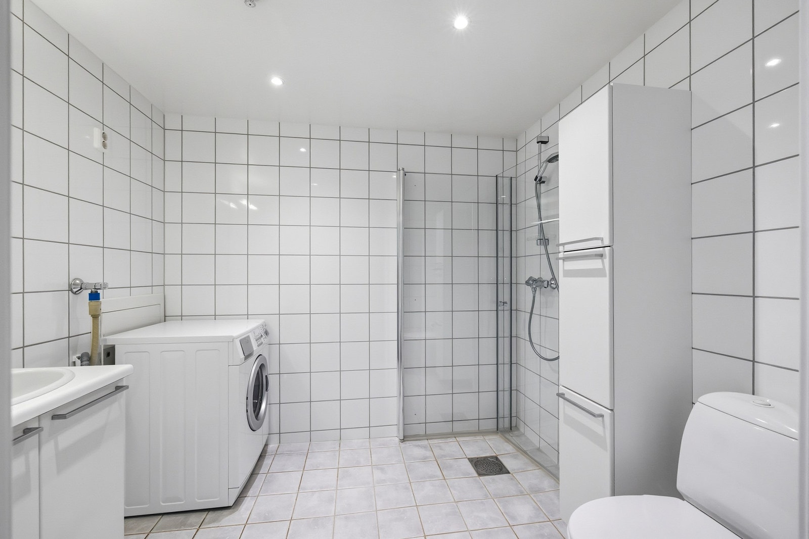 Flislagt bad med varmekabler i gulv og opplegg for vaskemaskin. Badet er innredet med dusjhjørnet med dører og vegghengt toalett. Galleribilde