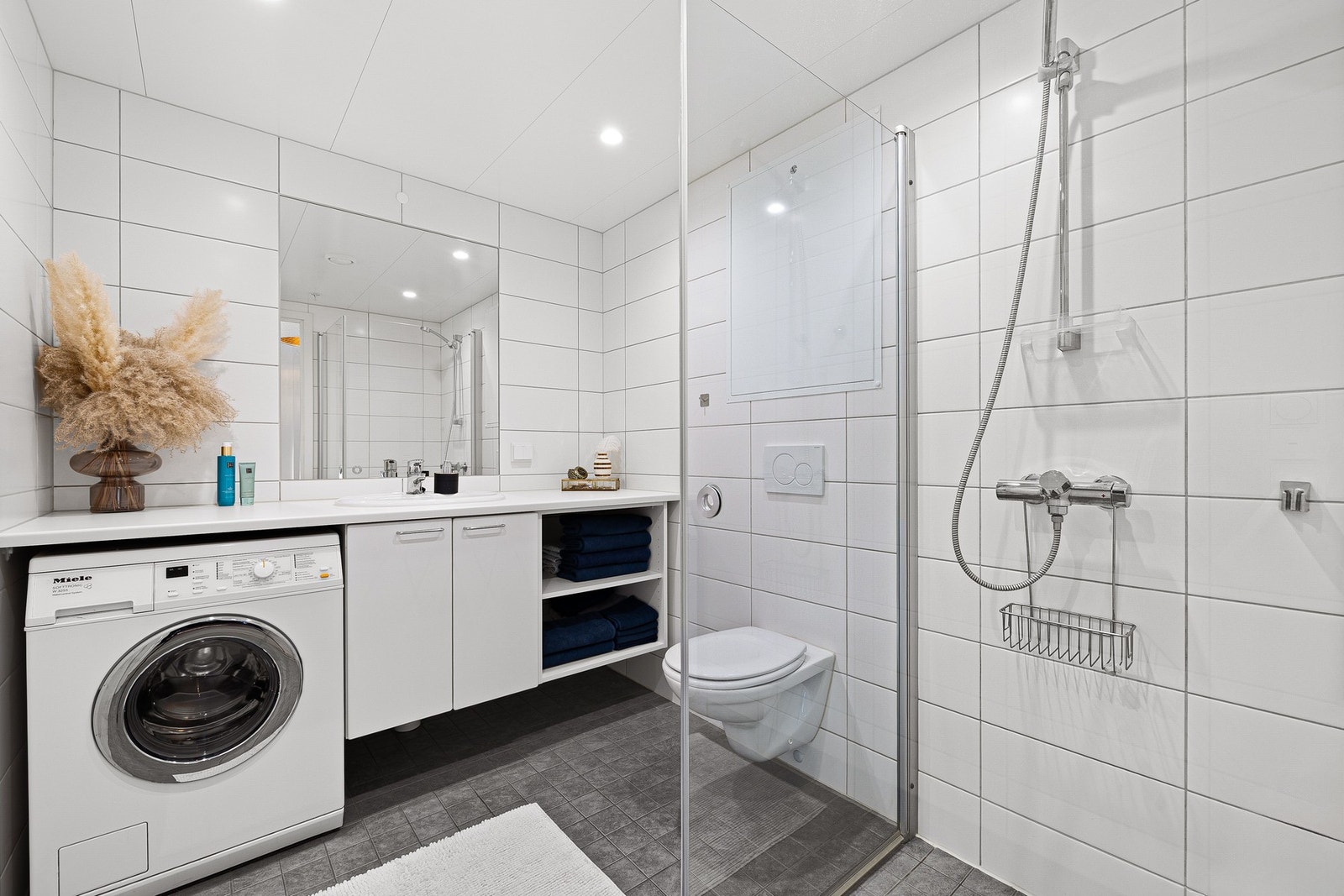 Badet fremstår innbydende og tidløst. Gråe gulvfliser med elektriske varmekabler, flis på vegg og plater i himling. Legg merke til den praktiske servantplaten som strekker seg fra vegg til vegg. Galleribilde