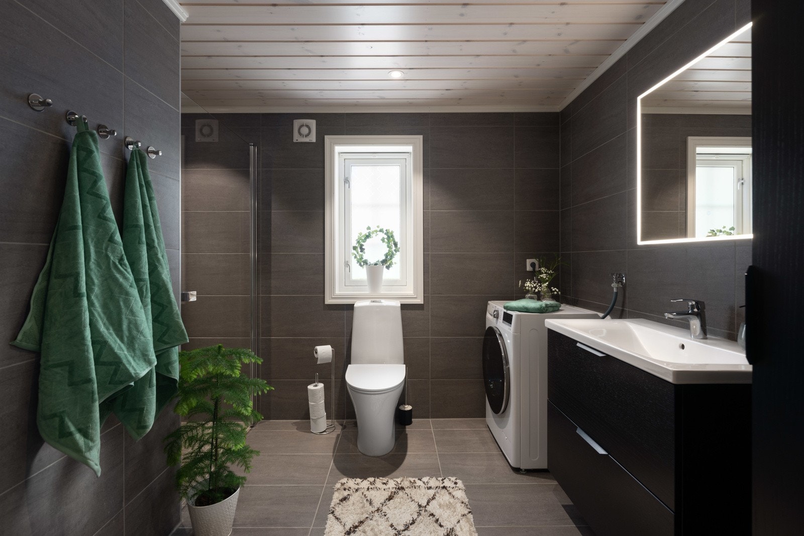 Stort moderne bad med fliser fra gulv til tak med varmekabler i gulv. Baderommet er innredet med helstøpt servantplate med skuffer i svart. over er det satt inn et moderne speil med lys rundt. Galleribilde