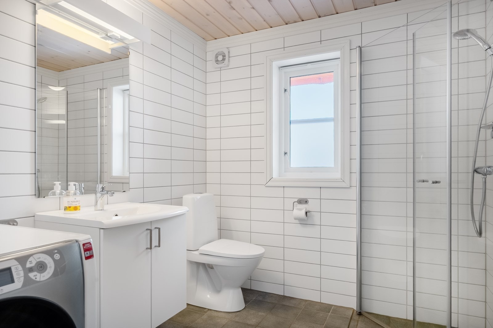 Pent baderom med lyse fliser på vegg og grå fliser på gulv. Badet har gulvvarme og opplegg for vaskemaskin. Galleribilde