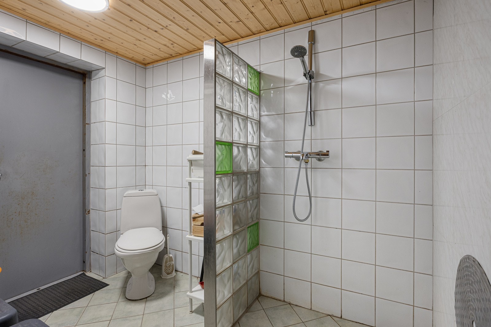 Badet i underetasjen har direkte adkomst fra garasjen og hvite fliser og plater på veggene, samt lyse fliser på gulvet. I tillegg til toalett har badet et dusjhjørne med glassbyggerstein. Galleribilde