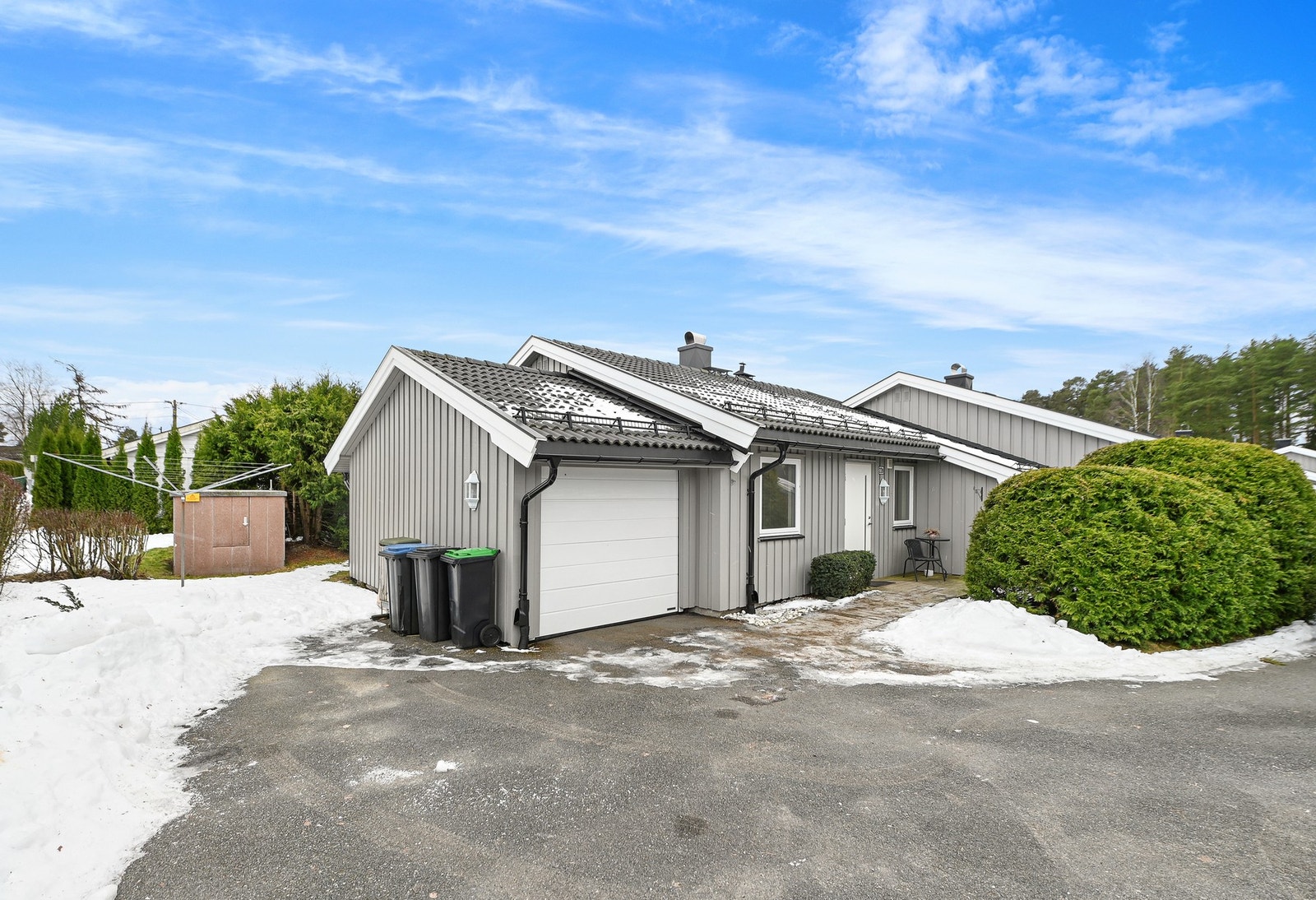 Grå enebolig med garasje, asfaltert innkjørsel og vinterhage. Snødekt uteområde med busker og trær. Klar blå himmel. Galleribilde