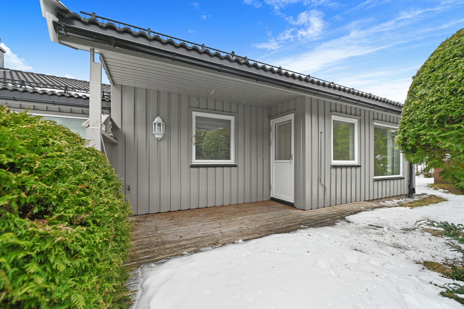 Grått hus med utendørs tredekke og snø på bakken. Huset har hvite vinduskarmer og en hvit ytterdør. Galleribilde