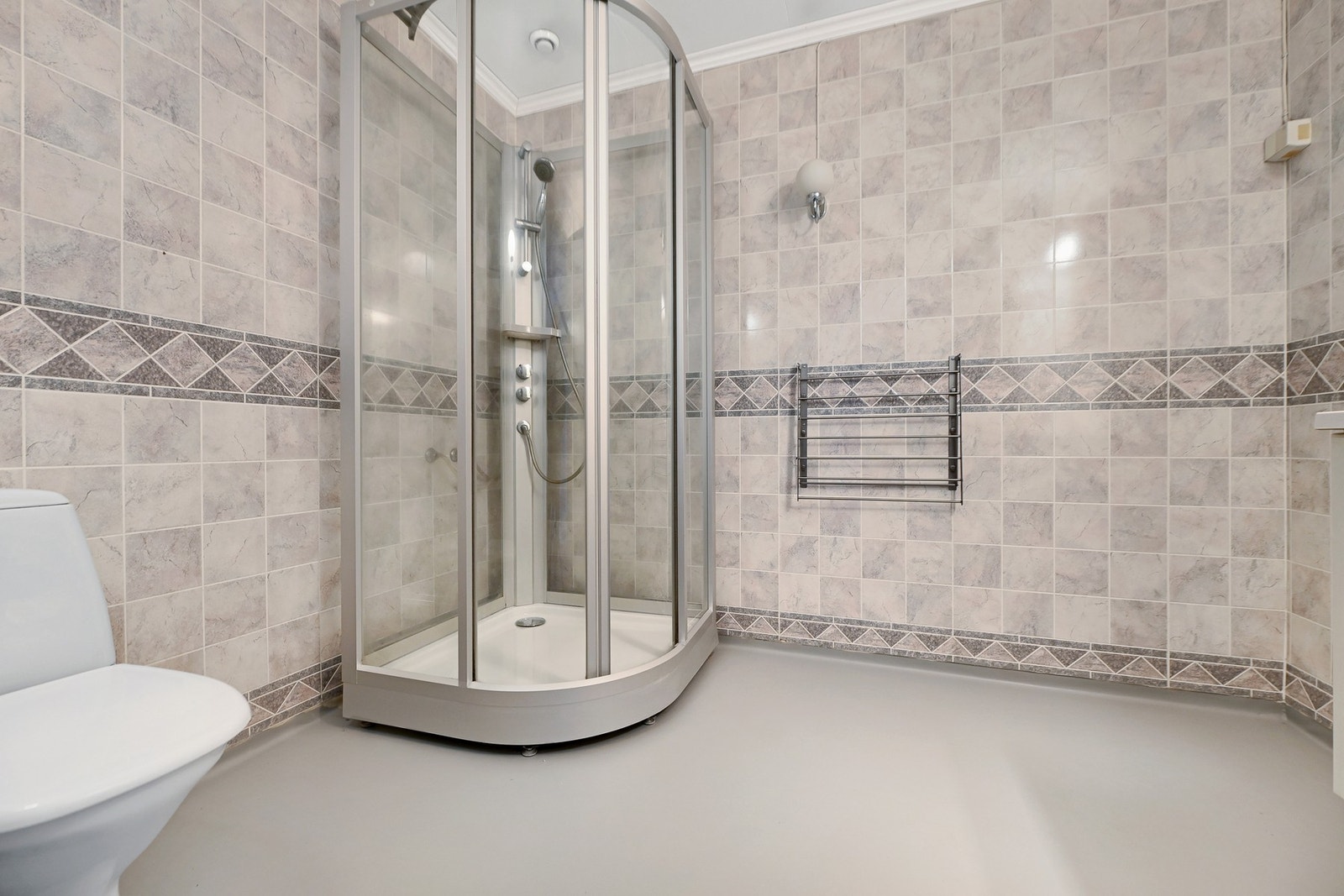 Badet har dusjkabinett, toalett og flislagte vegger med dekorative detaljer. Vegghengt håndkleholder gir ekstra komfort. Galleribilde