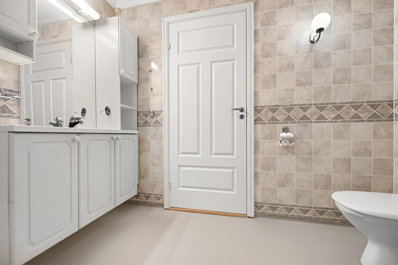 Flislagt bad med lyse vegger, speilskap og servant med underskap. Vegghengt toalett og enkel belysning. Galleribilde