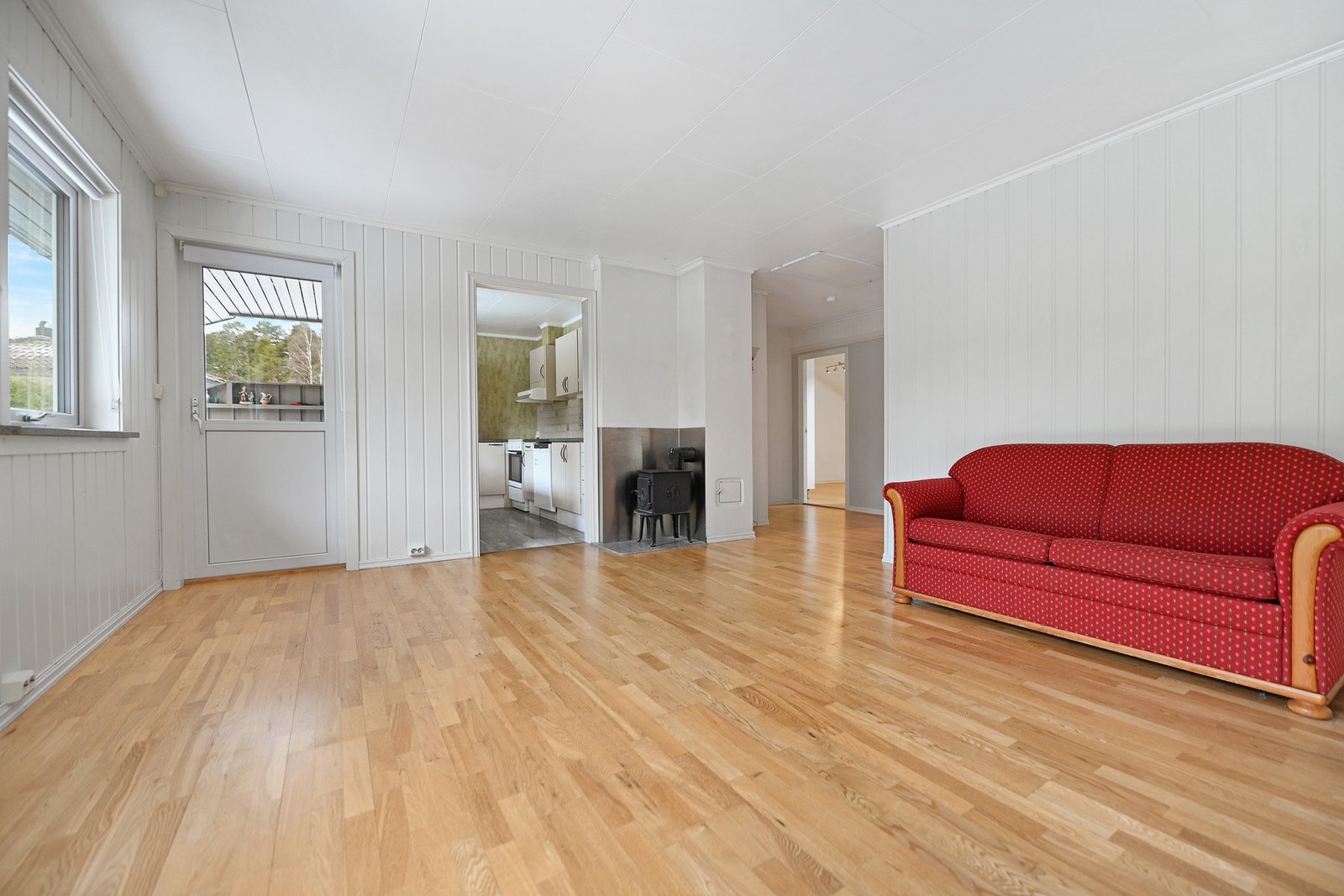 Lyst rom med parkettgulv, vedovn, og tilgang til kjøkken gjennom åpen dør. Galleribilde