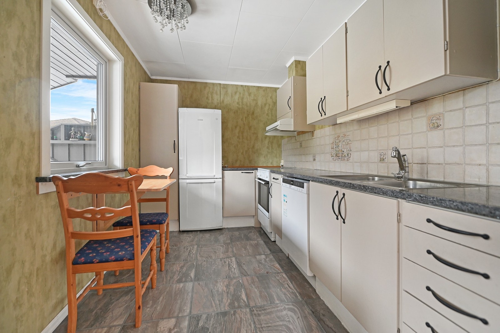 Kjøkken med lyse skap og hvitevarer, flislagt vegg, stort vindu og spiseplass. Galleribilde