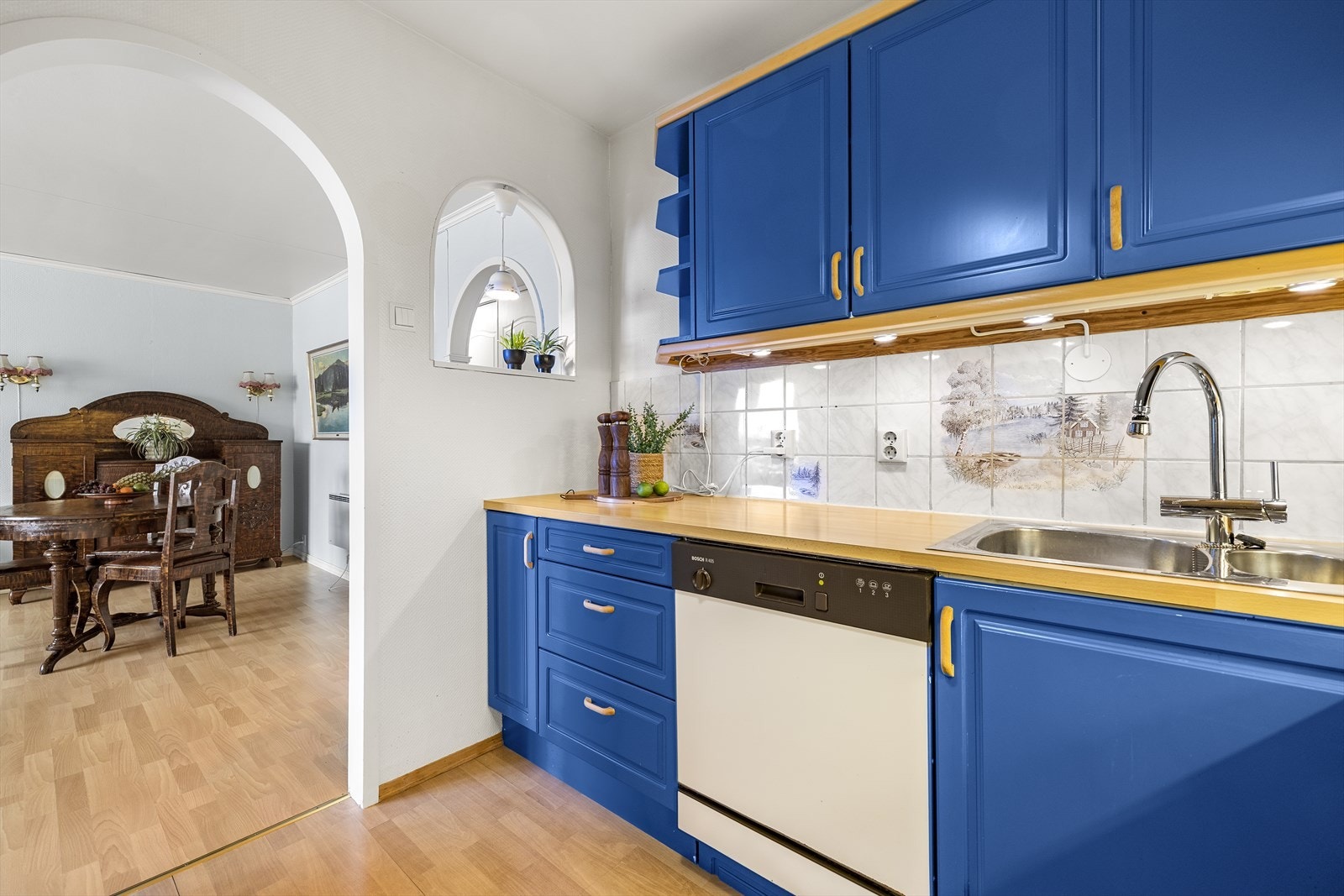 Fliser med dekorativt mønster mellom benkeplate og overskap. Det er plass til oppvaskmaskin, komfyr og frittstående kjøleskap i innredningen. Galleribilde