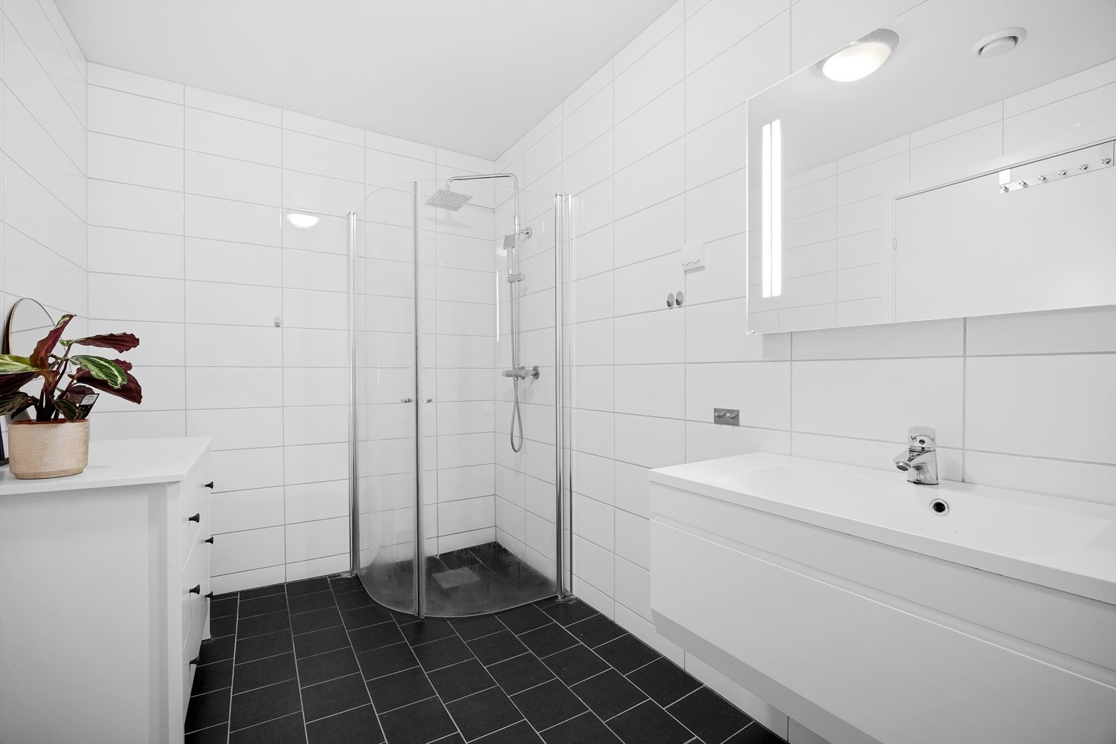 Badet er helfliset og innredet med dusjhjørne, vegghengt toalett og nedfelt porselen vask i pent baderomsmøbelement. Galleribilde