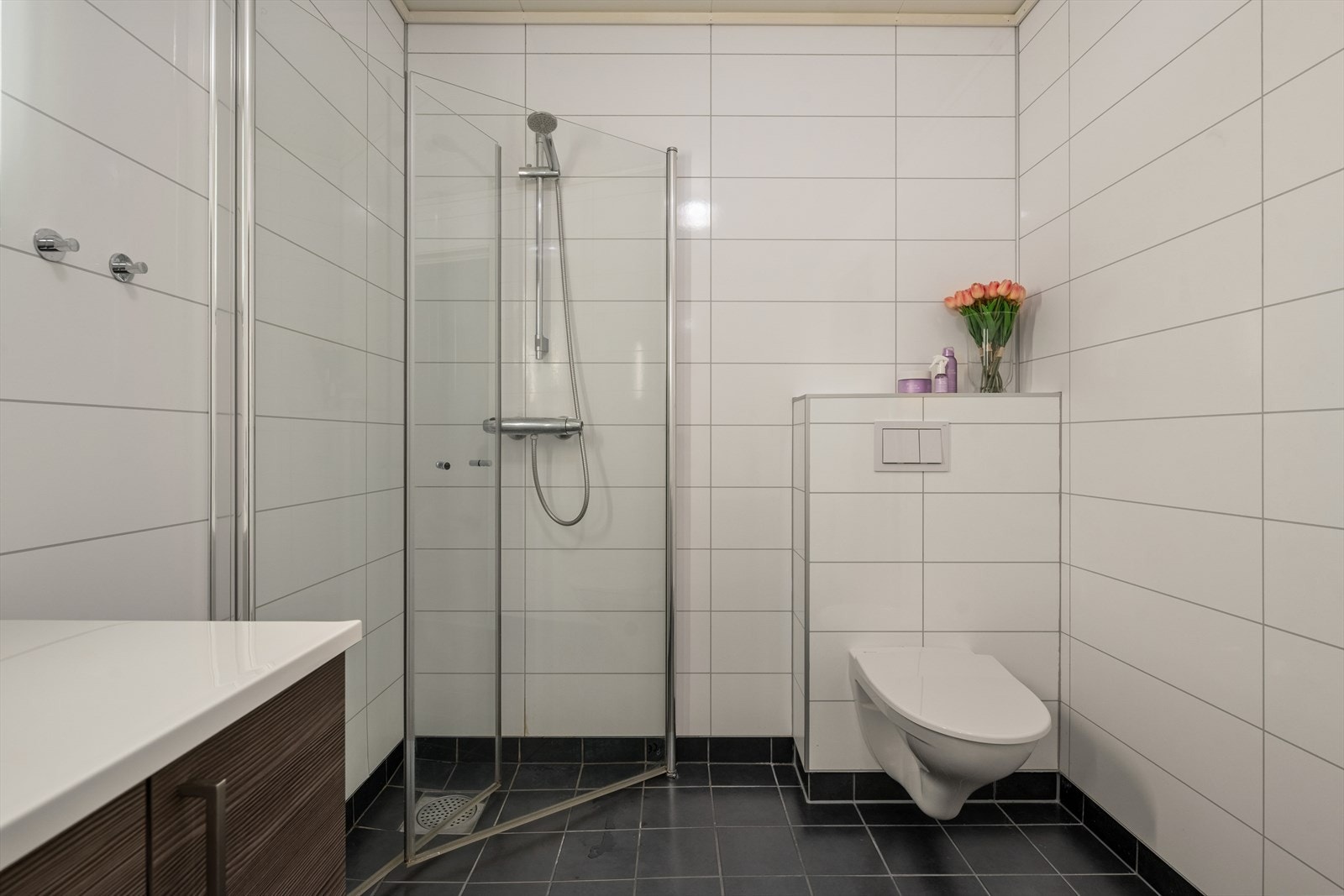 Baderommet er utstyrt med en baderomsinnredning med heldekkende servant, oppbevaring i underskap, speil, veggmontert toalett og et dusjhjørne med foldbare dusjdører Galleribilde