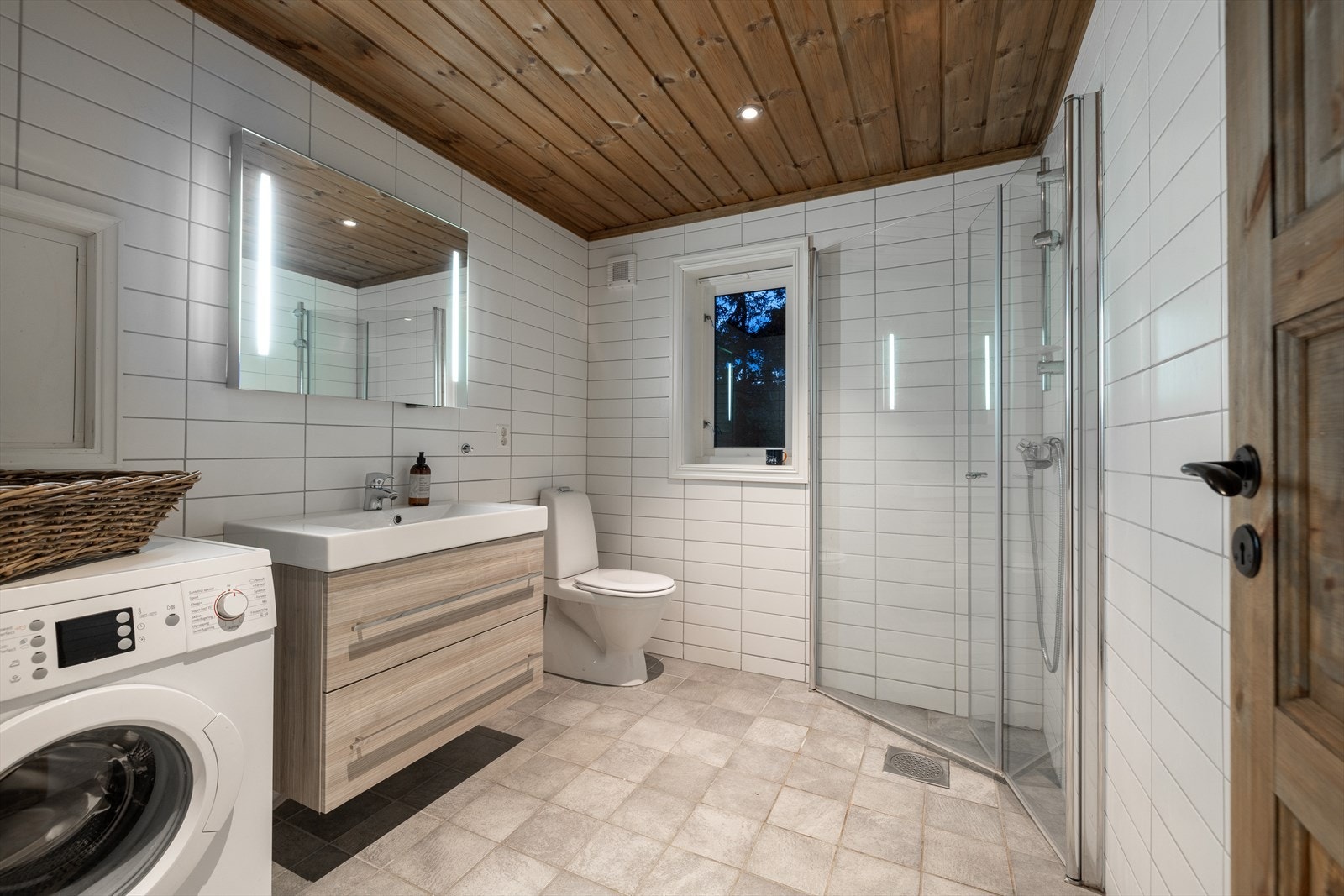 Pent helfliset baderom av romslig størrelse, innredet med dusjhjørne med innfellbare dusjvegger, toalett, servantskap og vaskemaskin Galleribilde