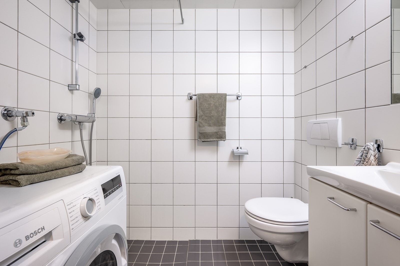 Badet er belagt med lyse fliser på vegger og mørkere kontrastfliser på gulv. Det er lagt varmekabler i gulv. Galleribilde