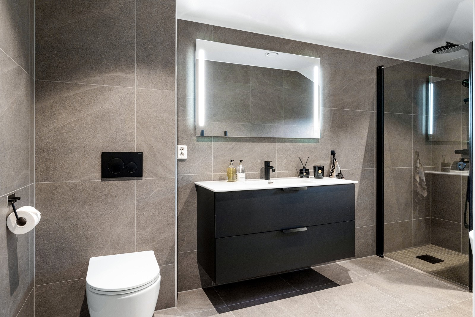 Badet er romslig og flislagt, og inneholder vegghengt toalett, servantinnredning, dusjhjørne og et innbydende badekar Galleribilde