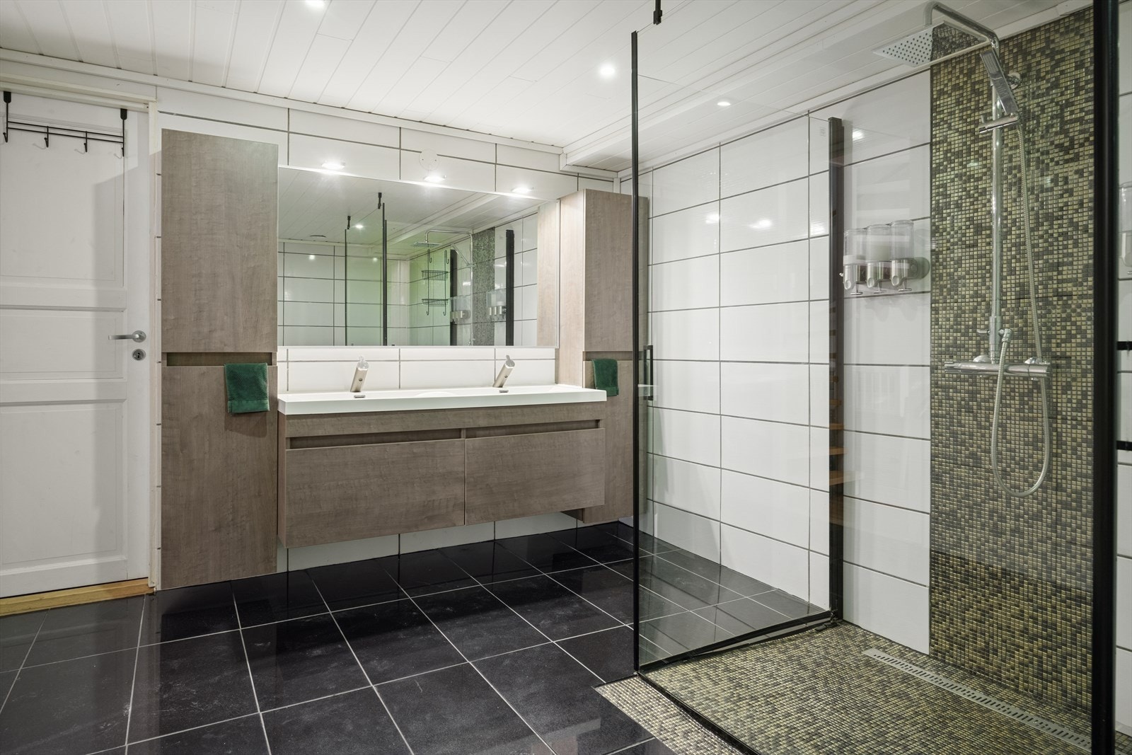 Innredet med dusjvegger av glass og dusjgarnityr på vegg, vegghengt toalett, innredning med servant og opplegg for vaskemaskin Galleribilde