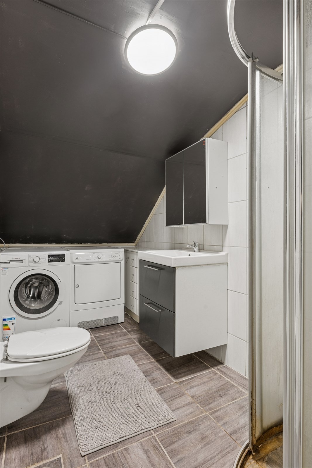 I tillegg har boligen et baderom i 2. etasje med flislagte overflater. Innredningen kombinerer glatte, profilerte og speilfronter. Her finner du også opplegg for vaskemaskin, som gjør baderommet enda mer funksjonelt. Galleribilde