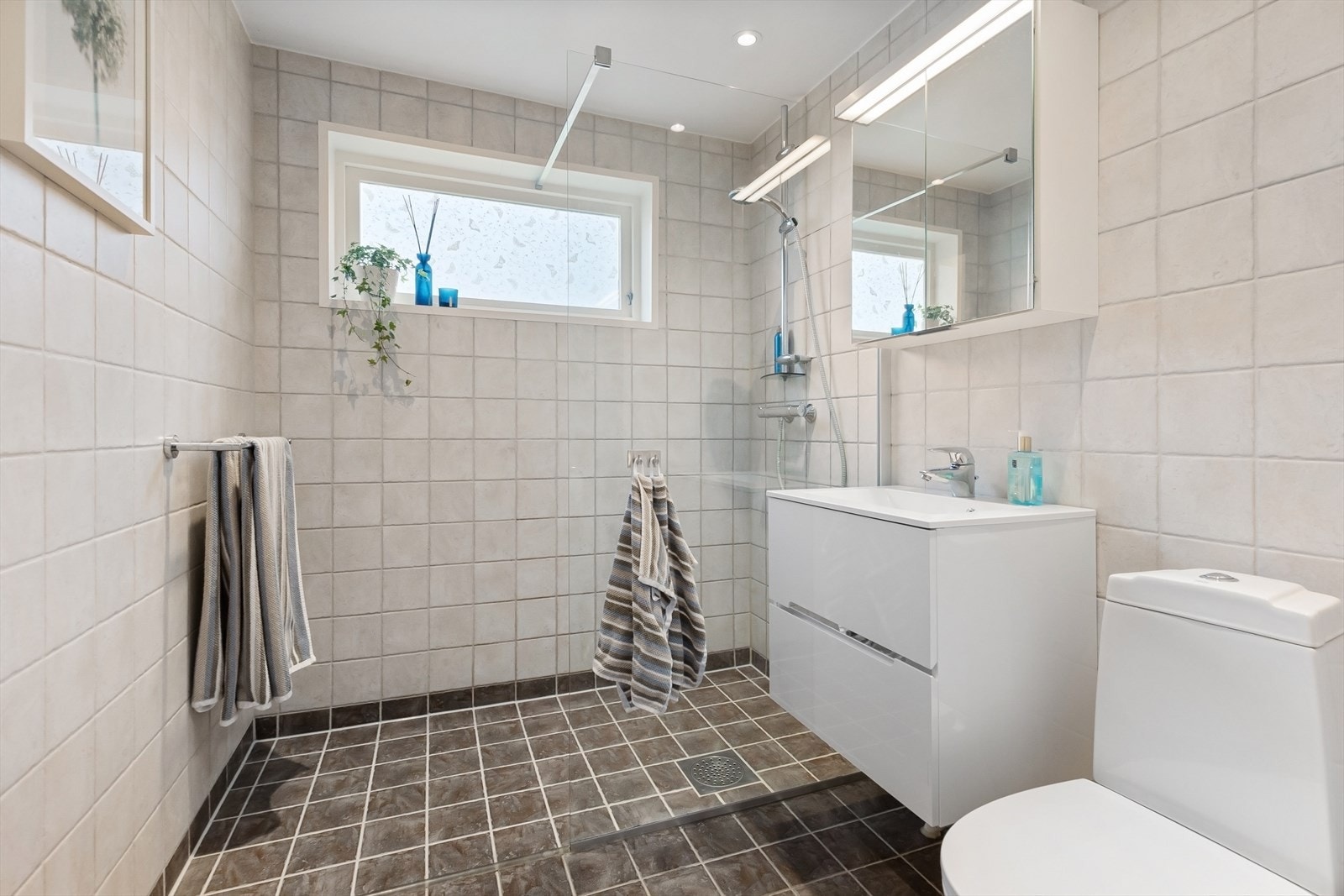 Lyst og pent bad i 1. etasje, med fliser på vegger og gulv. Badet er innredet med servant i innredning med to skuffer, speilskap med belysning, dusj med glassvegg og toalett Galleribilde
