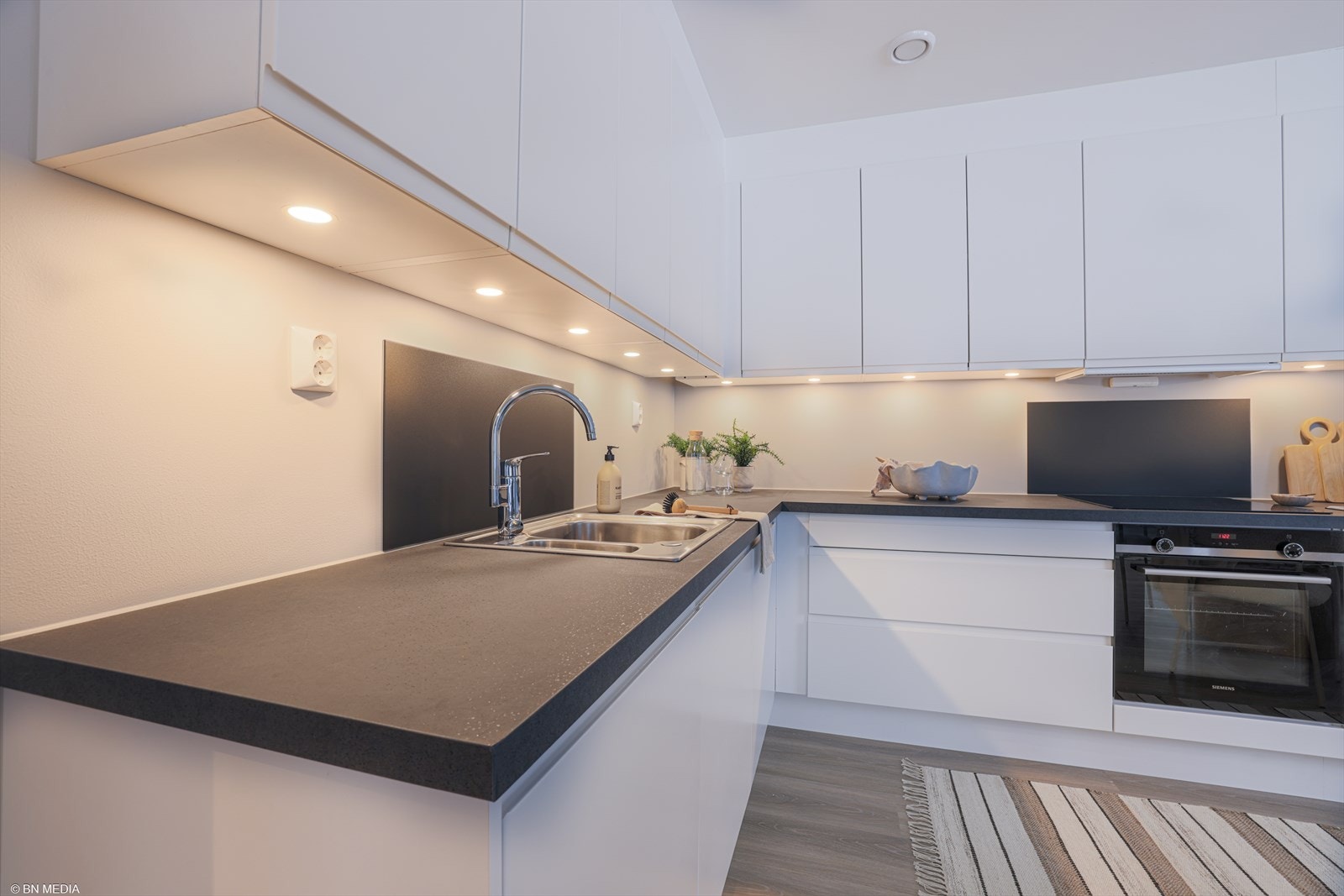 Kjøkkenet har en stilren Sigdal-innredning med hvite, glatte fronter og en mørk laminert benkeplate med nedfelt oppvaskkum i rustfritt stål. Galleribilde