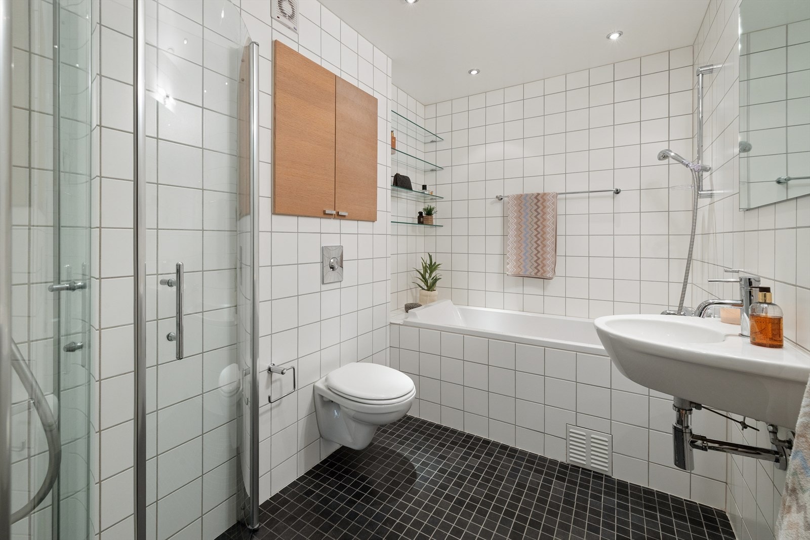 Badet er praktisk innredet med servant med innredning, veggmontert toalett, badekar og dusjhjørne med dusjvegger. Galleribilde