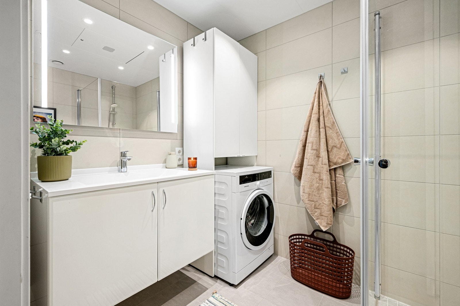 Flislagt og pent bad med praktiske innfellbare dusjdører i glass, dusjbatteri, avløp for vaskemaskin og veggmontert toalet Galleribilde