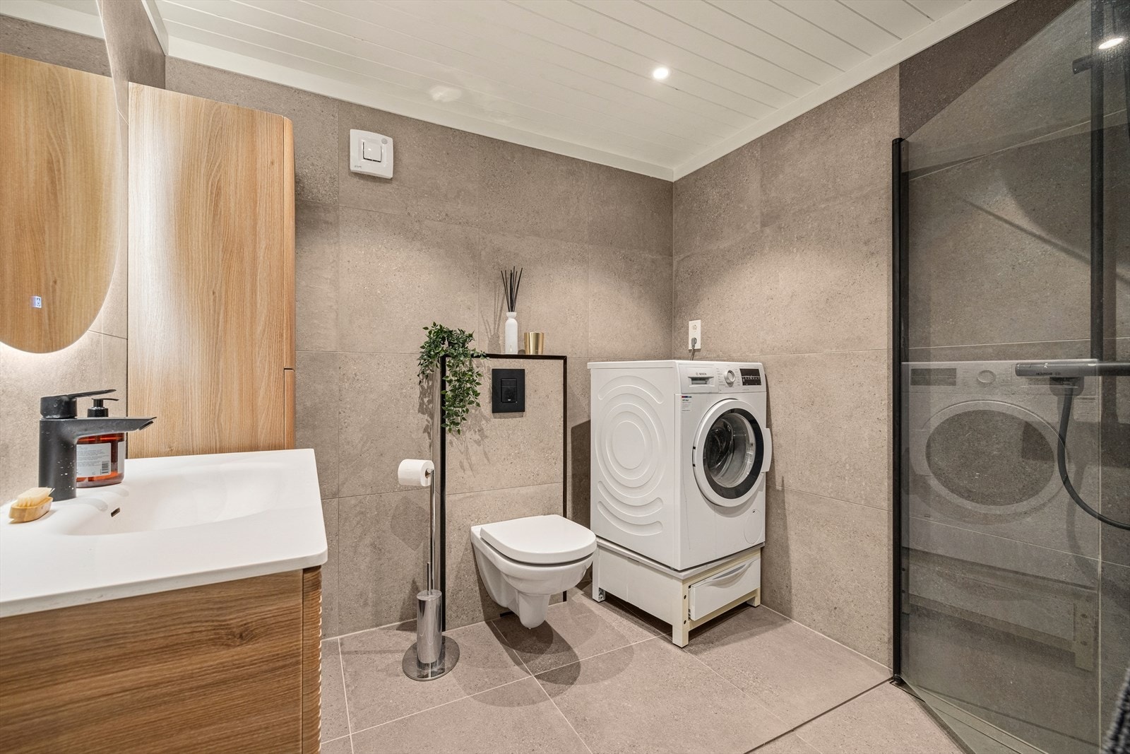 Innredning med nedfelt servant, veggmontert toalett, dusjvegger/hjørne og opplegg for vaskemaskin. Det er slukrenne ved vegg i dusjsone. Galleribilde