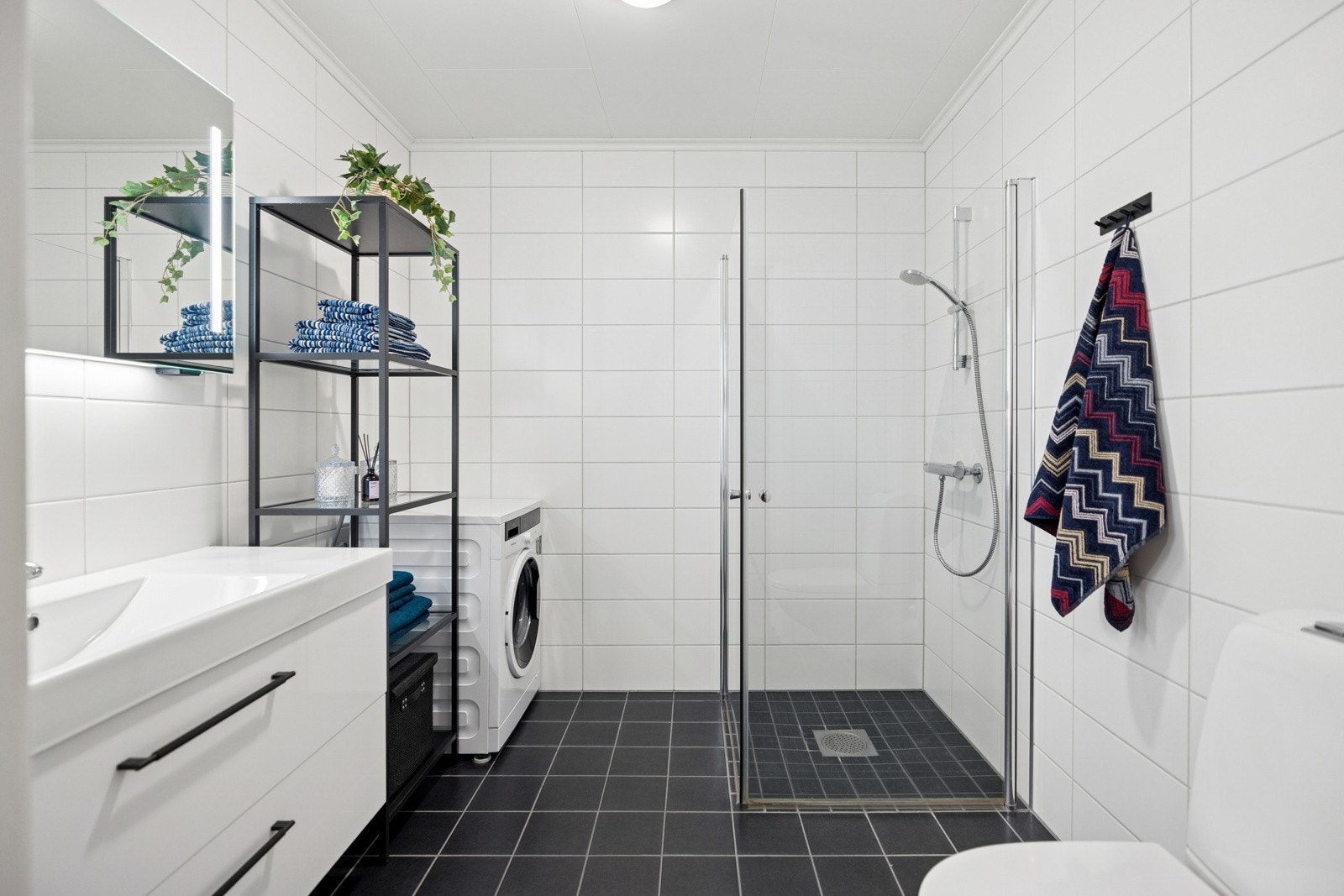 Videre har badet dusjnisje med glassdører som kan skyves mot dusjsone, wc og opplegg for vaskemaskin. Varme i gulvet bidrar til et behagelig rom for velvære Galleribilde