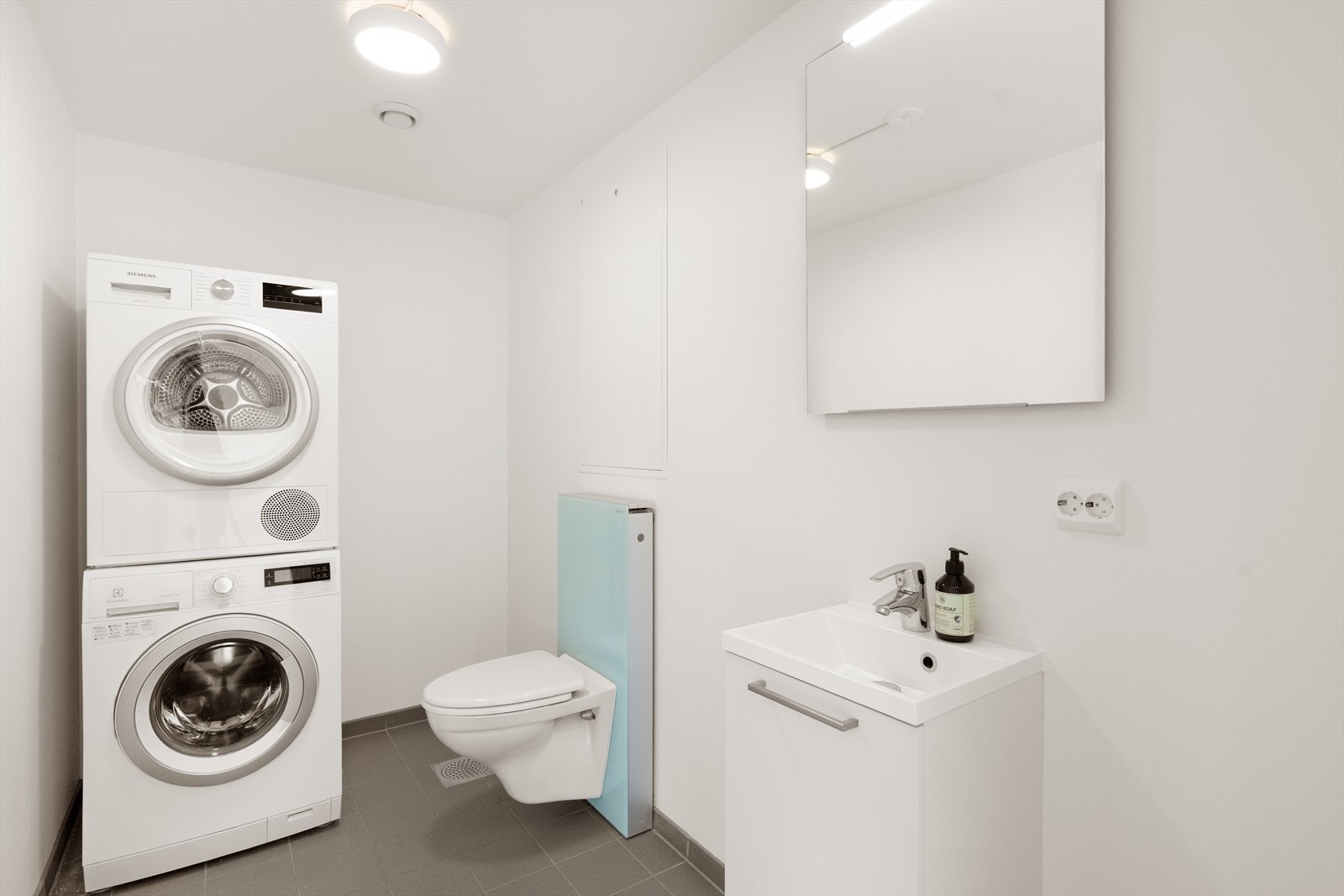 Praktisk og delikat vaskerom kombinert med toalettrom. Inneholder vegghengt toalett, innredning med servant, speil med lys, opplegg for vaskemaskin og tørketrommel. Galleribilde