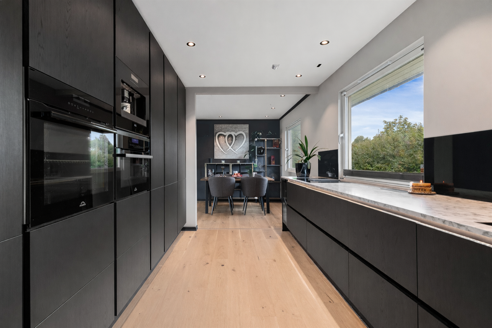 Innredet i mørke slette fronter, med en lekker benkeplate. Integrerte hvitevarer som mikro, stekeovn, kaffemaskin, kjøleskap og platetopp med ventilator medfølger. Redigert i chatgpt Galleribilde