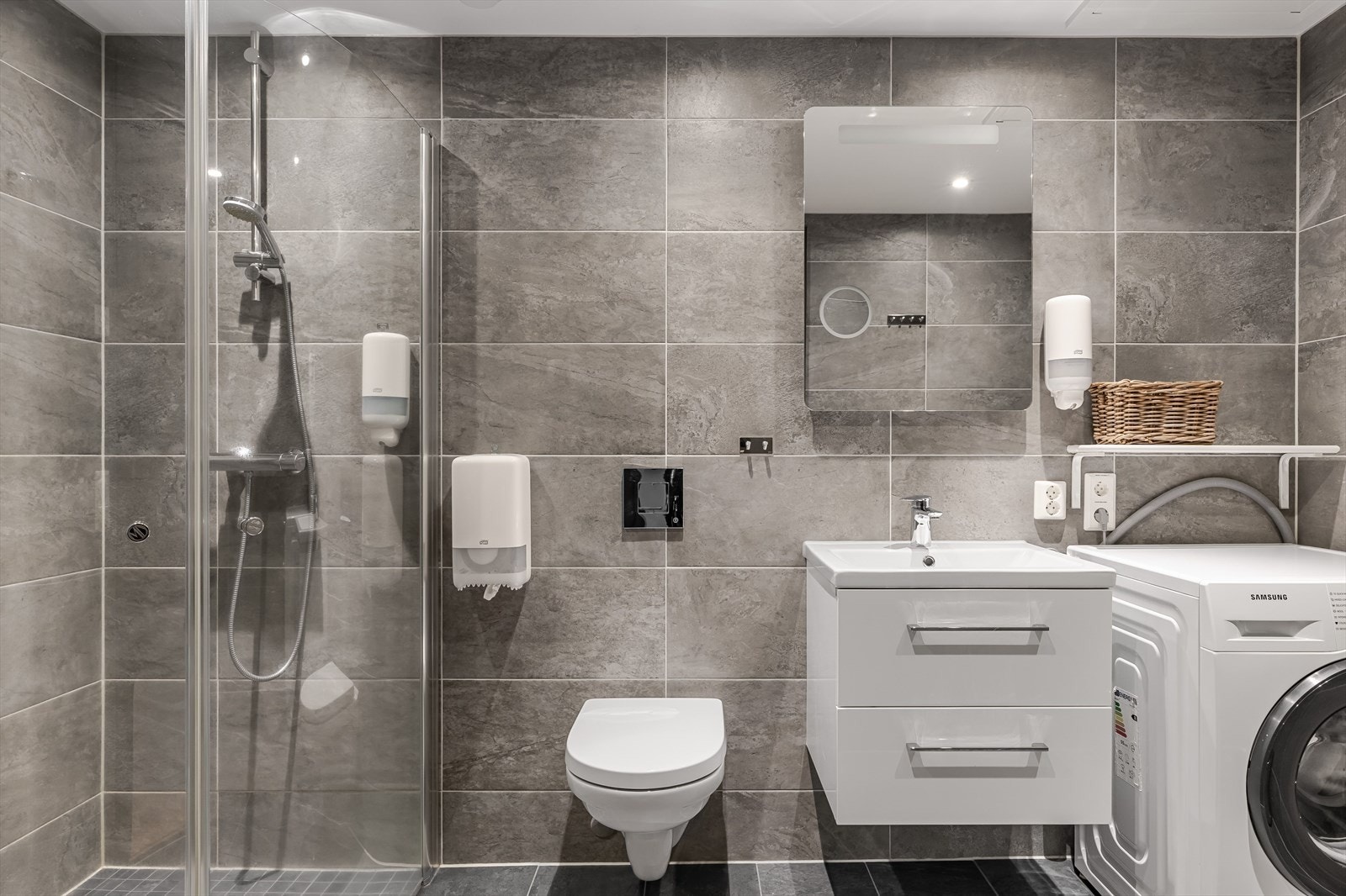 Moderne baderomsinnredning med skuffer og ovenpåliggende servant, speil med integrert belysning over. Baderommet er også utstyrt med dusjhjørne med innfellbare glassdører og vegghengt toalett. Galleribilde