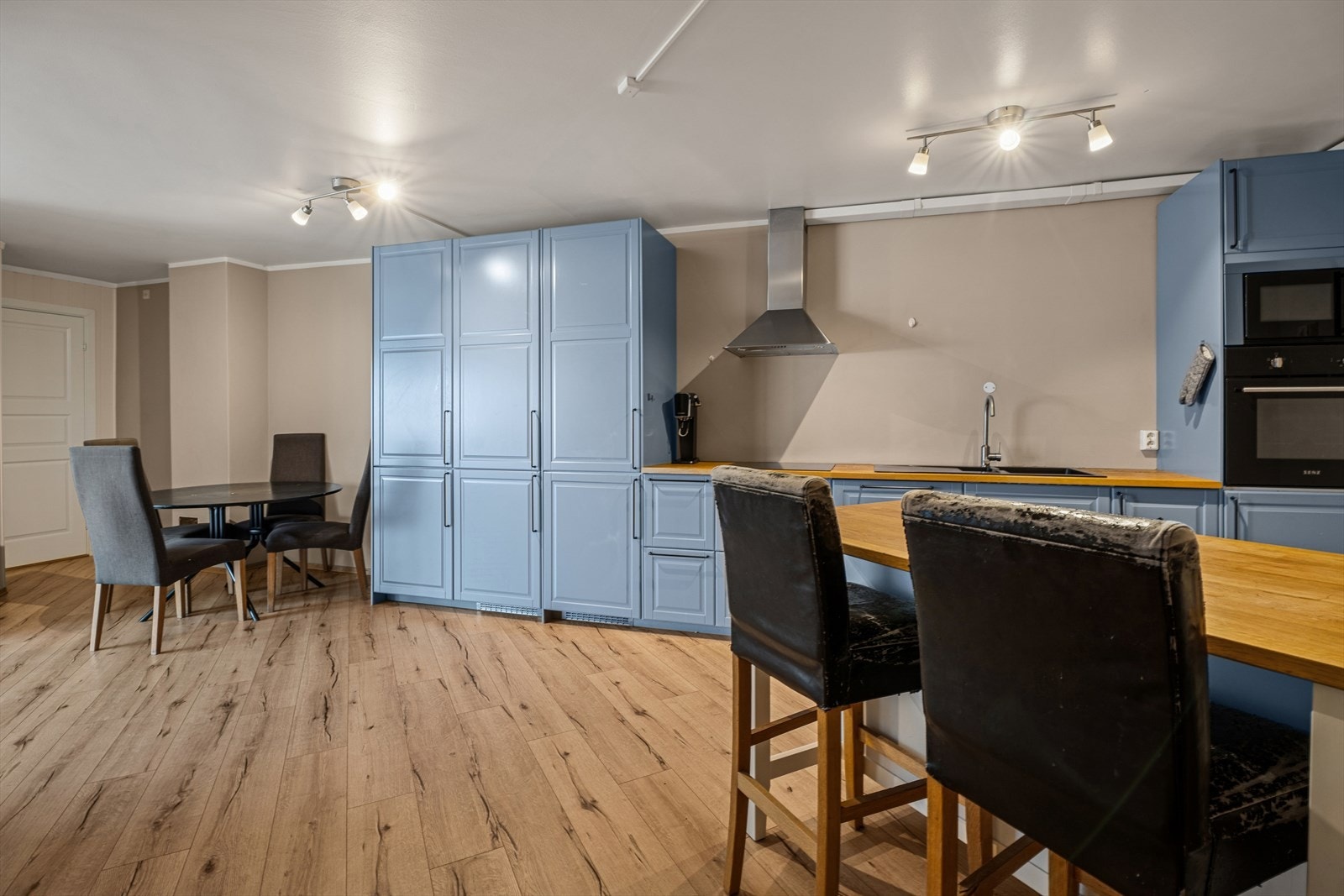 Benkeplaten i heltre tilfører en varm og naturlig finish, og er utstyrt med nedfelt kjøkkenkum som gir en praktisk og elegant arbeidsflate. Over kokesonen er det montert en effektiv kjøkkenventilator som sørger for godt avtrekk under matlaging. Galleribilde