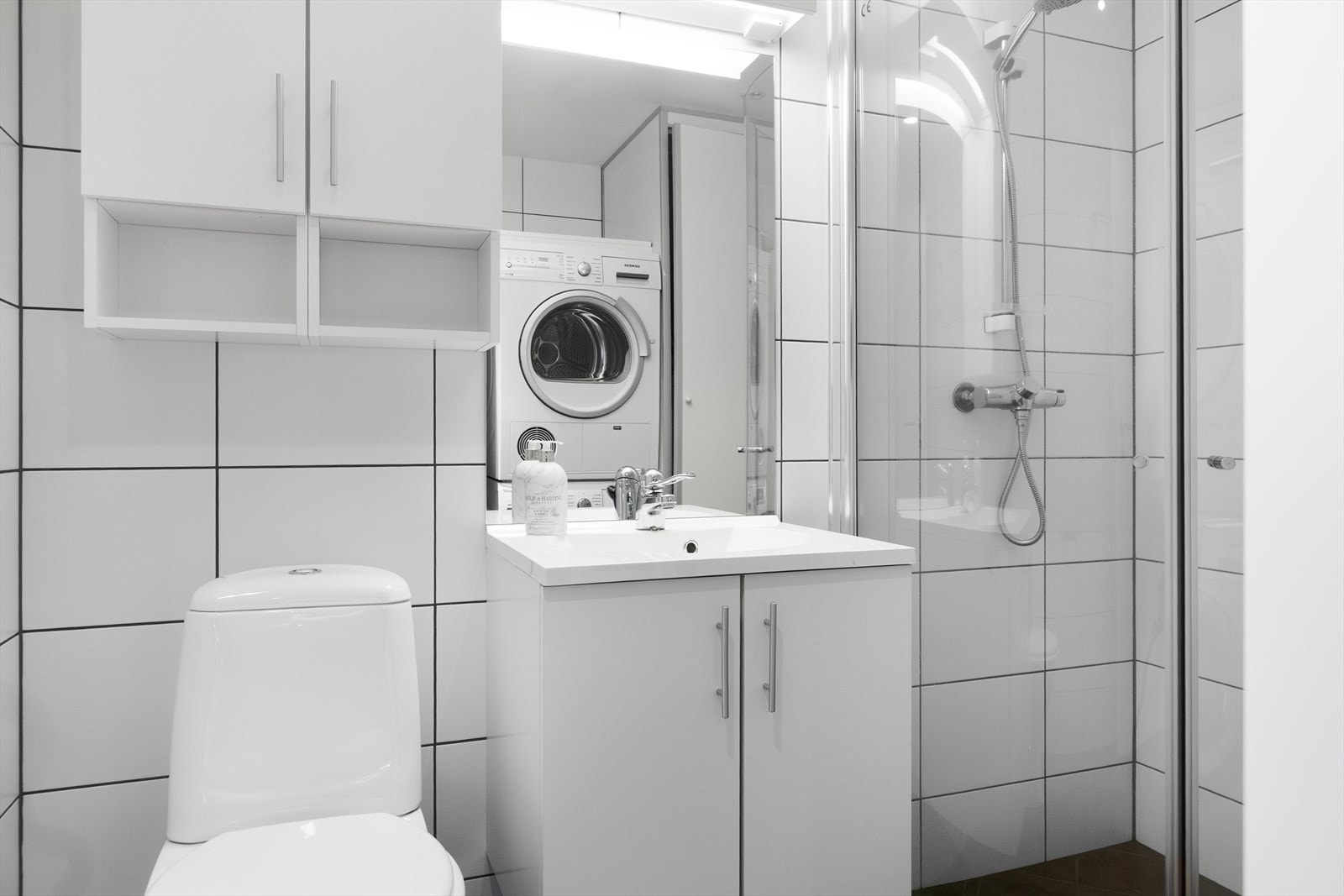 Badet er lyst og delikat med flislagte vegger og flislagt gulv med varmekabler. Her er det dusjhjørne med innfellbare glassdører, toalett og opplegg for vaskemaskin. Galleribilde