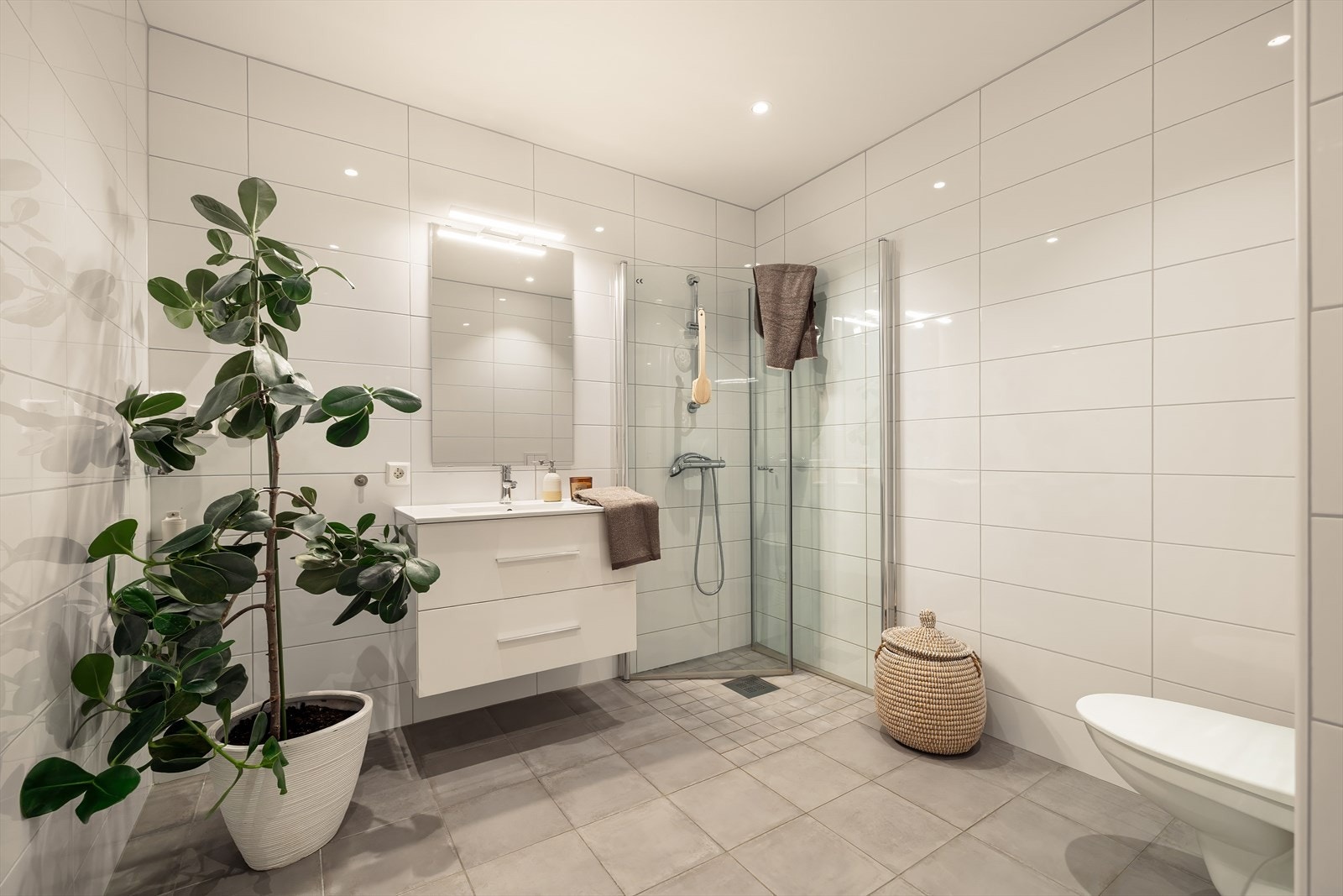 Hovedbaderommet har keramiske fliser på både gulv og vegger, og er funksjonelt innredet med vegghengt toalett, dusj med glass foldedører, servant med møblement, speil og belysning. Galleribilde