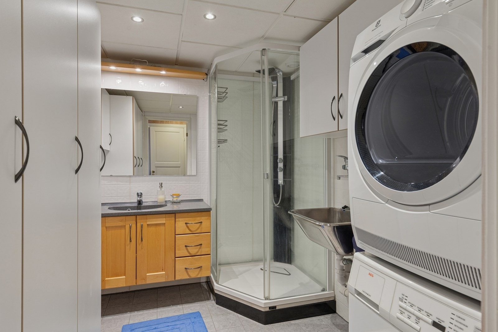 Bad/vaskerom med dusjkabinett, opplegg for vaskemaskin, utslagsvask og baderomsinnredning. Galleribilde